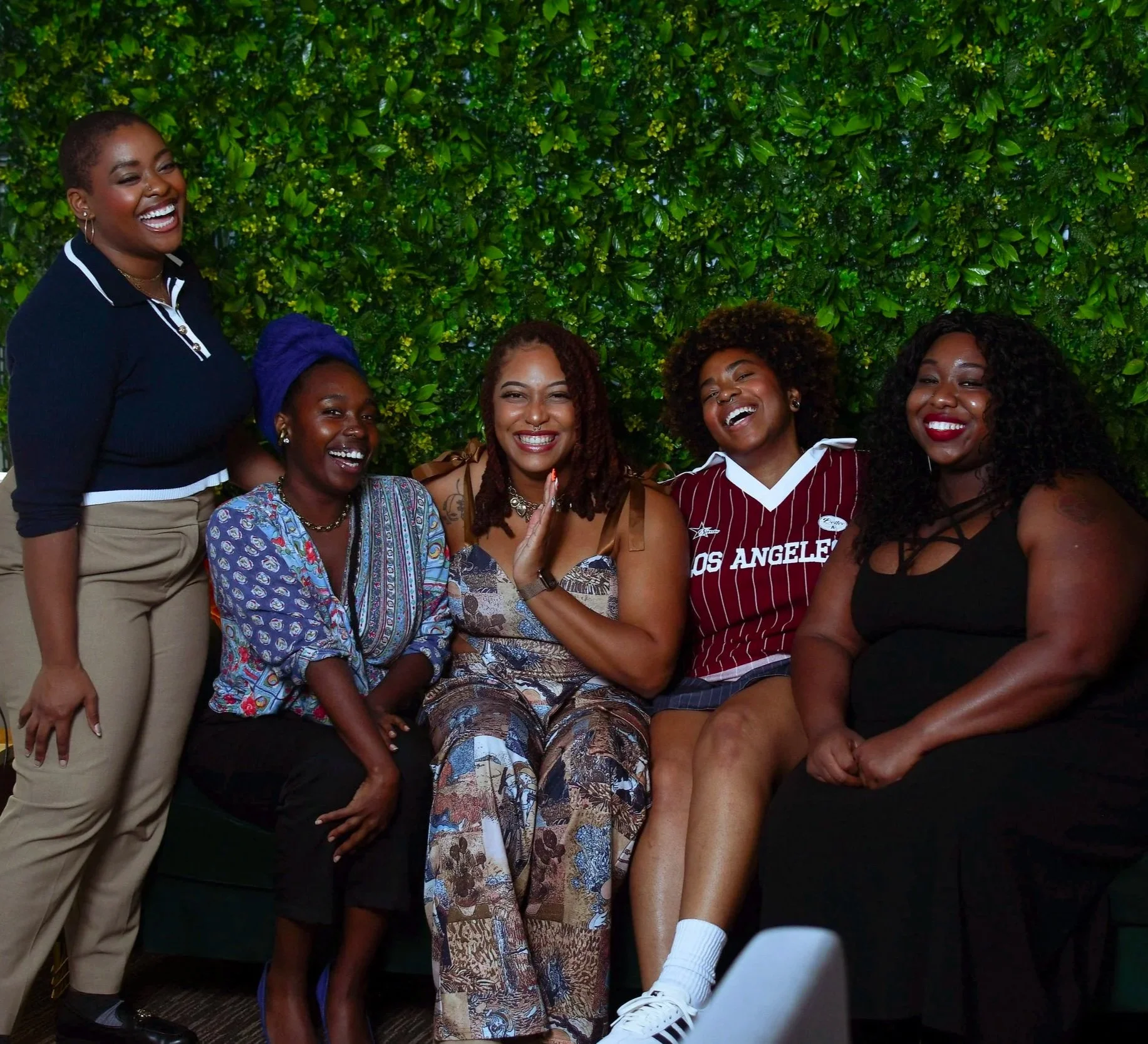 Group of seven women smiling and laughing together indoors in front of a green wall.