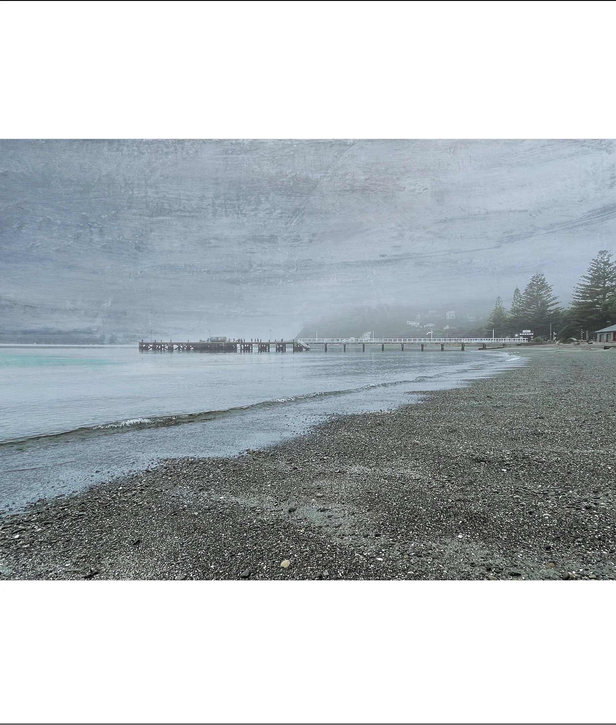 View of a shoreline with small waves on a pebble beach, a pier extending into the water, and a distant city clouded by fog or smoke, with trees along the coastline.