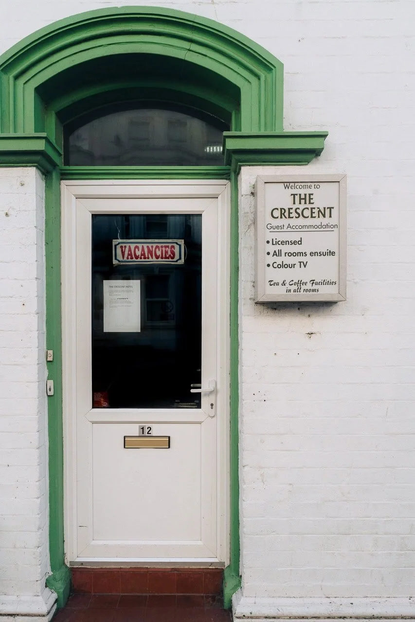 White door with the number 12, a 'VACANCES' sign in the window, and a sign beside the door welcoming to THE CRESCENT hotel, advertising licensed guest rooms, ensuite rooms, color TV, and tea and coffee facilities.