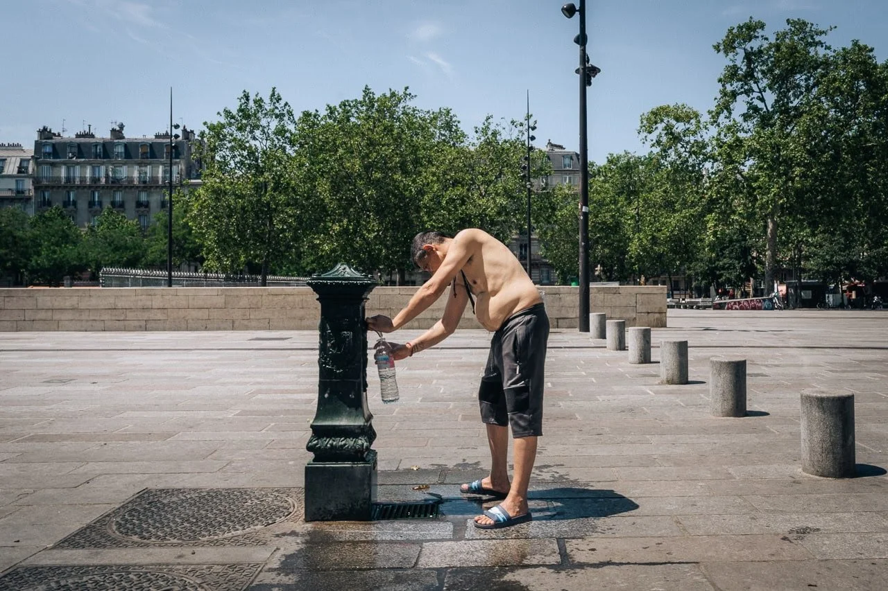 A shirtless man filling his water bottle at a public fountain in Paris, France