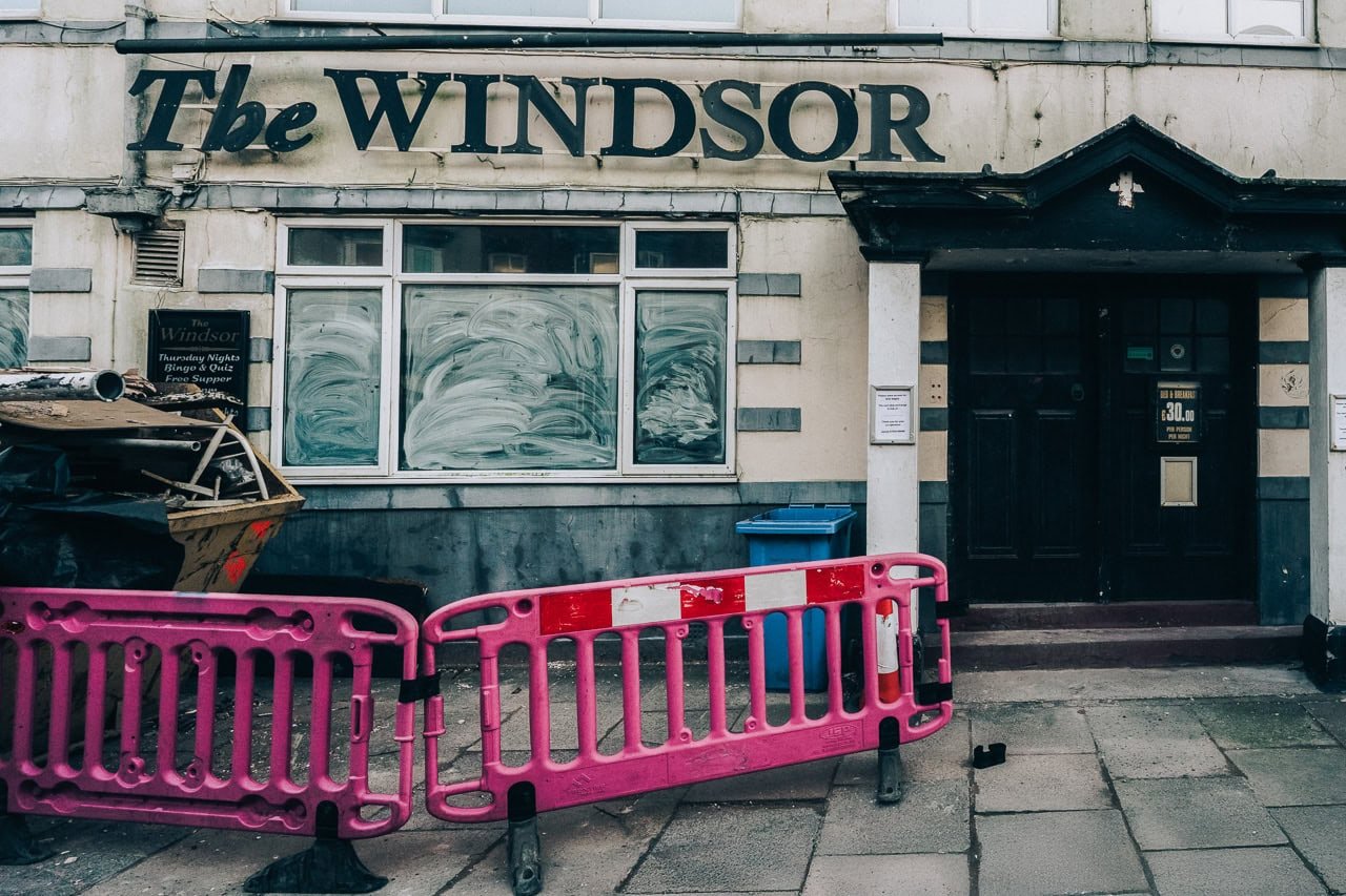 The Windsor pub exterior with boarded-up windows, a black door, and pink construction barriers in front.