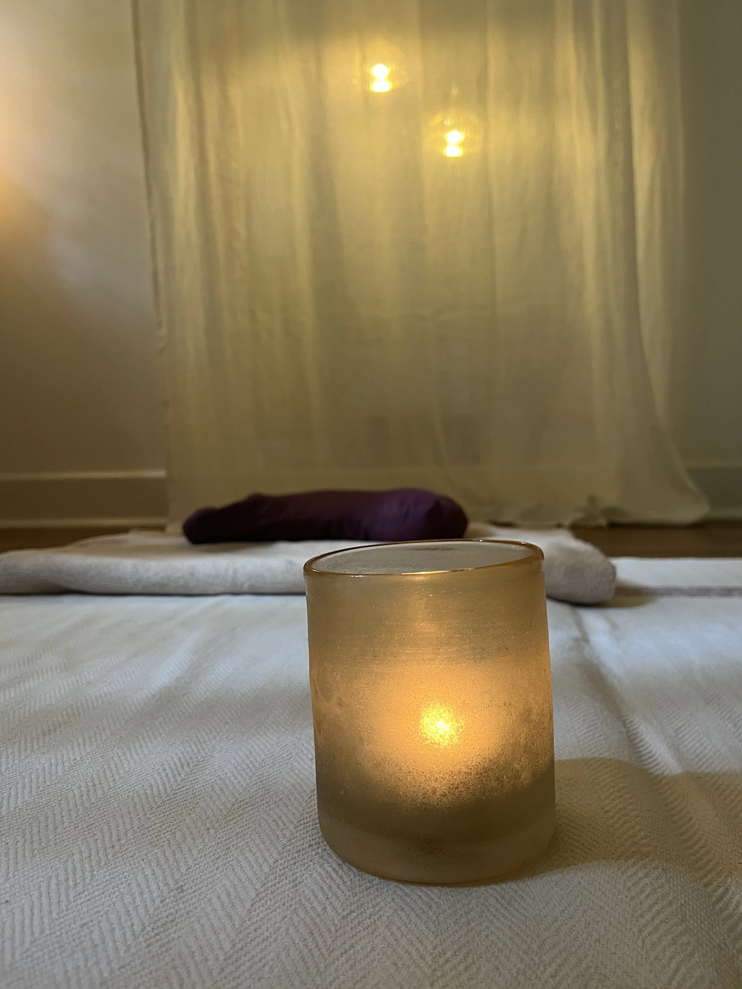 Kerze auf weißem Tuch in einem gemütlichen Raum, im Hintergrund ein Bett und Vorhänge mit warmem Licht.