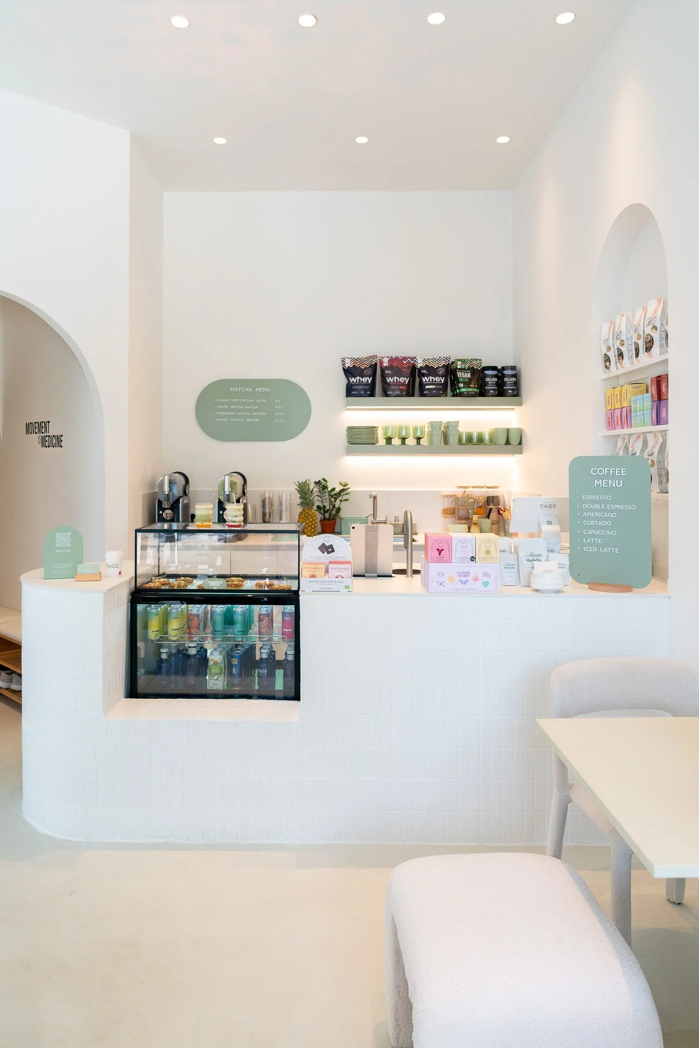 A minimalist white coffee shop counter with a display case of pastries, a small drink fridge, green coffee cups on shelves, and a coffee menu sign.