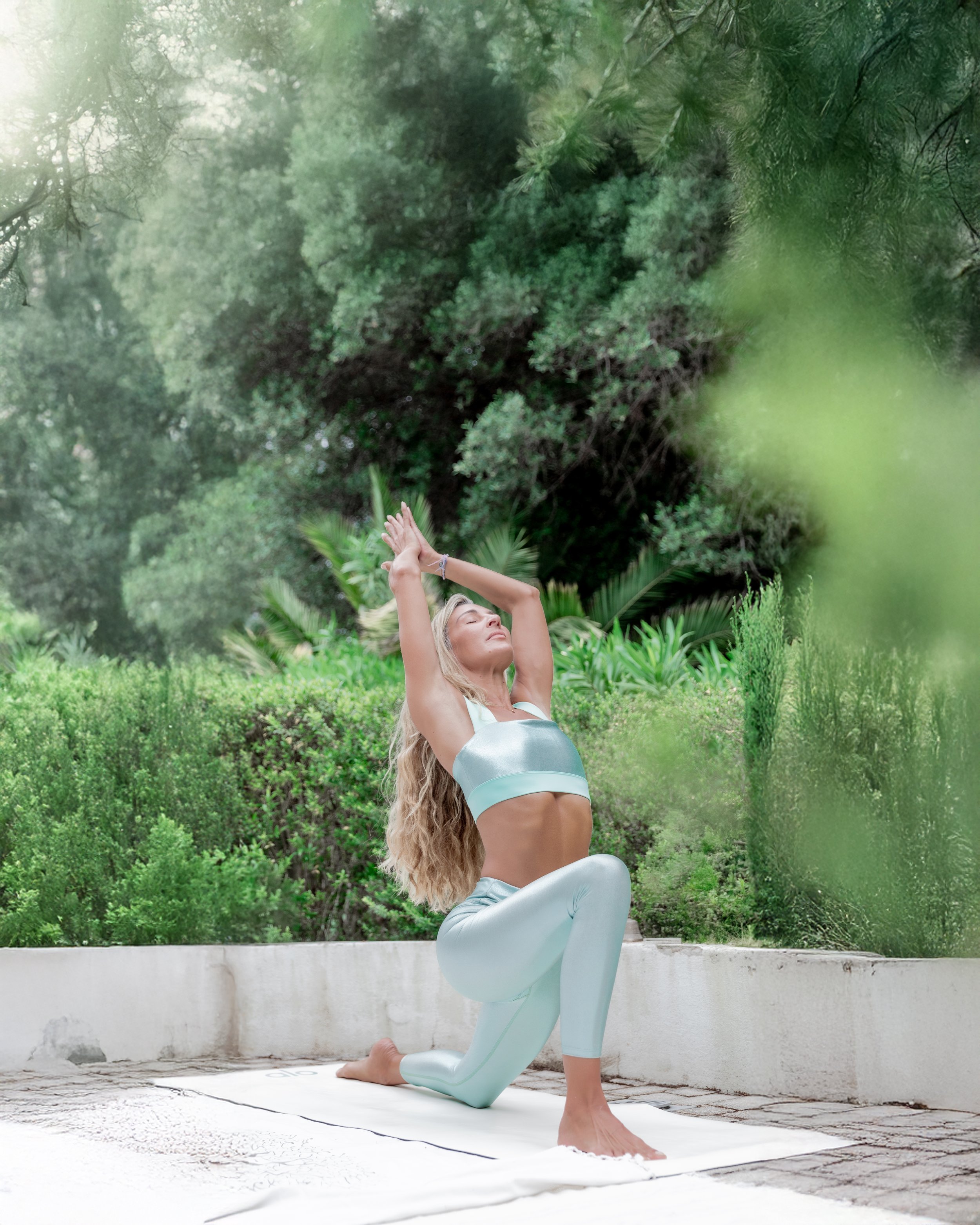 Woman practicing yoga outdoors on a white mat, kneeling with arms raised above her head, surrounded by lush green trees and plants.
