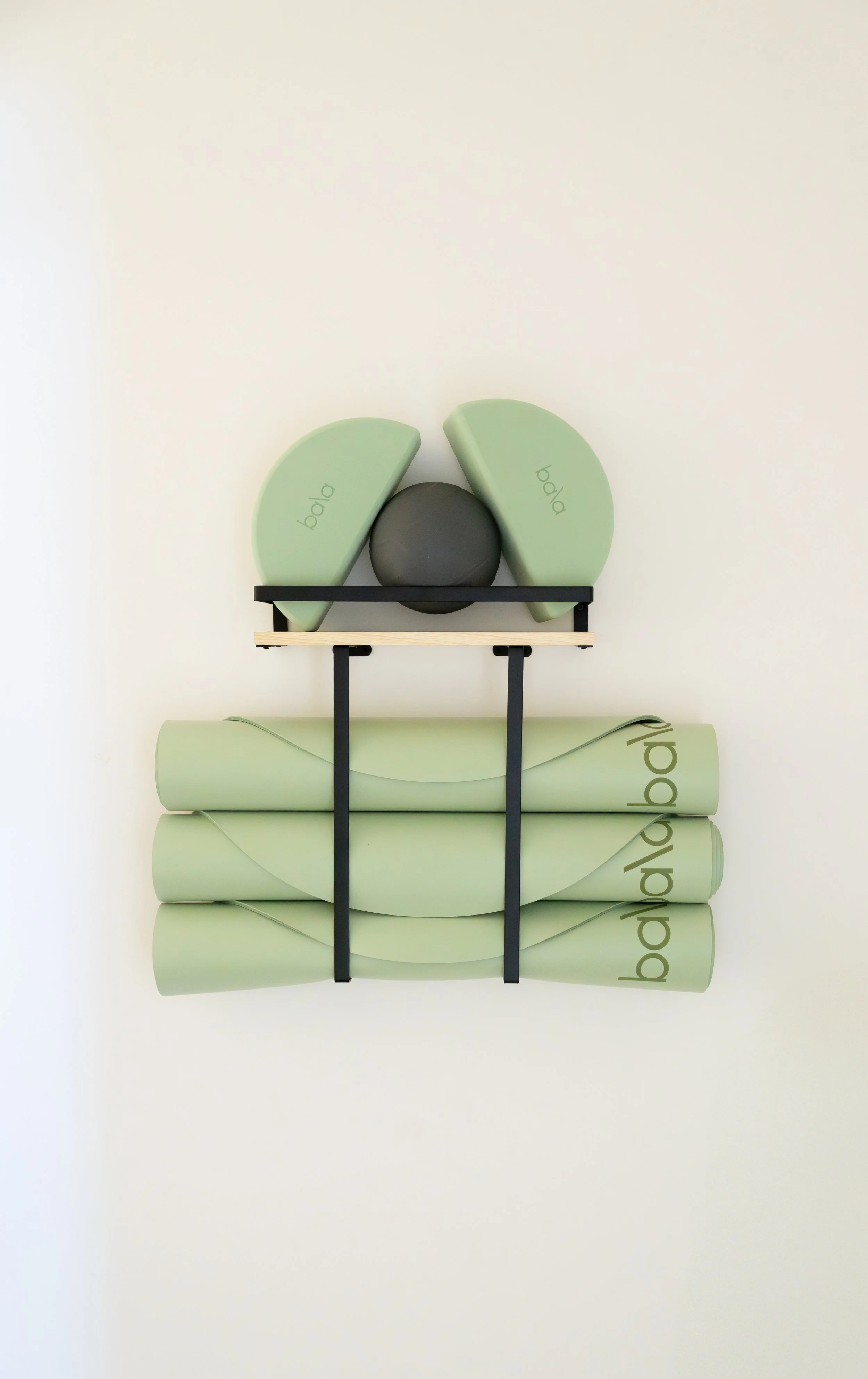 Stacked green yoga mats, black metal and wooden wall art piece with black and green foam blocks and a black ball on a white wall.
