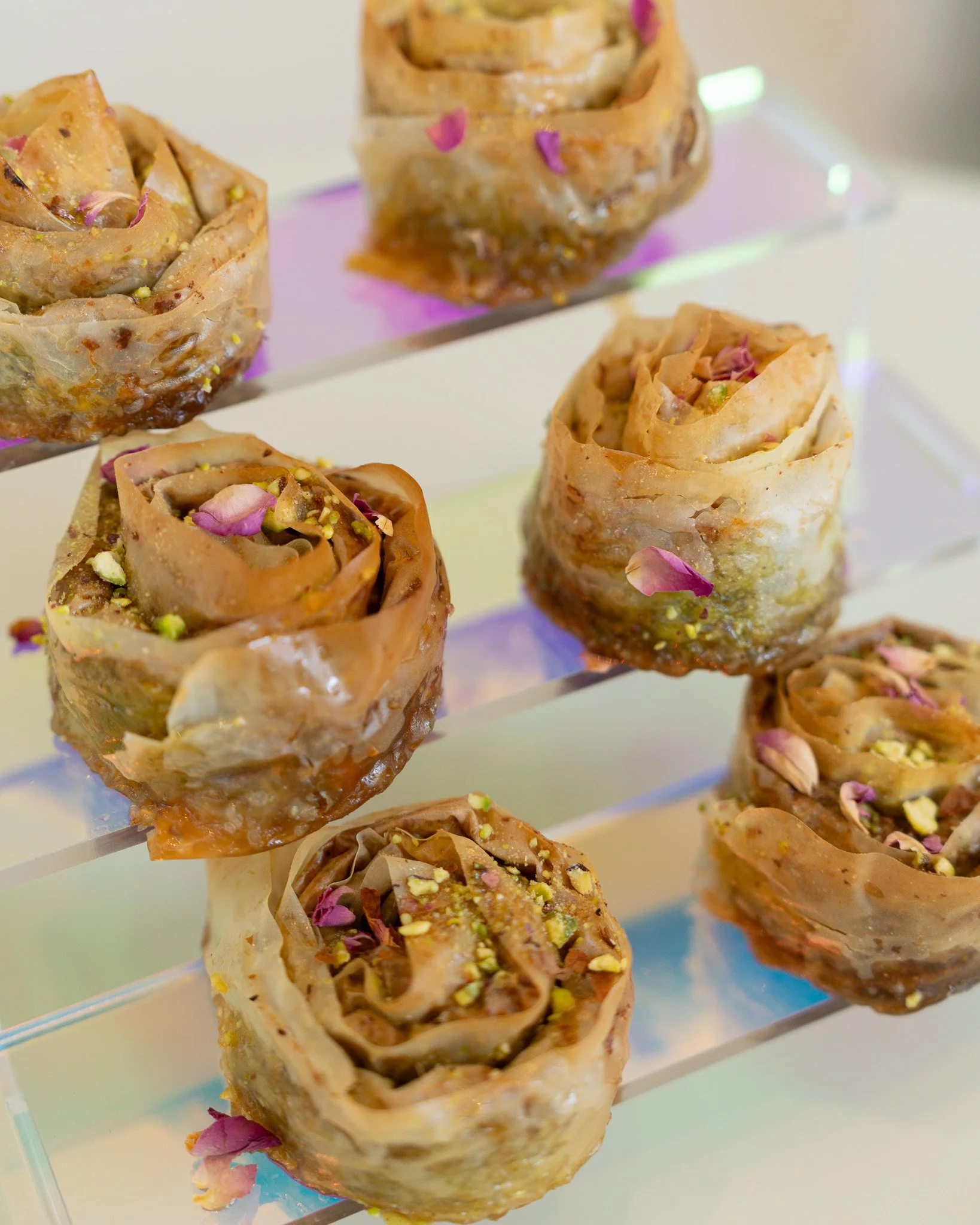 Close-up of baklava rolls topped with pink flower petals and crushed pistachios, arranged on a multi-layered acrylic stand.