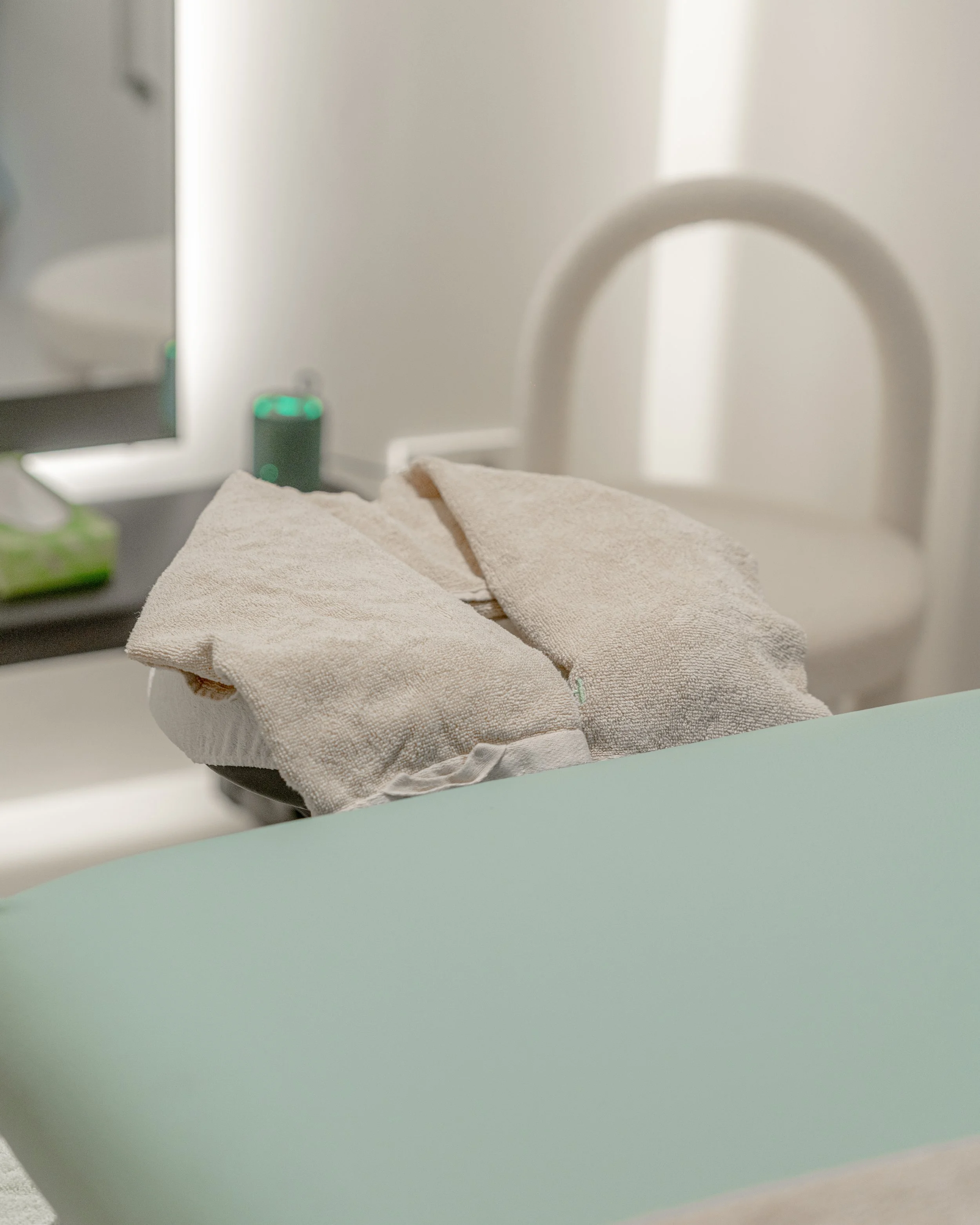A stool with rolled beige towels in front of a bathroom sink and mirror, with a toilet in the background.