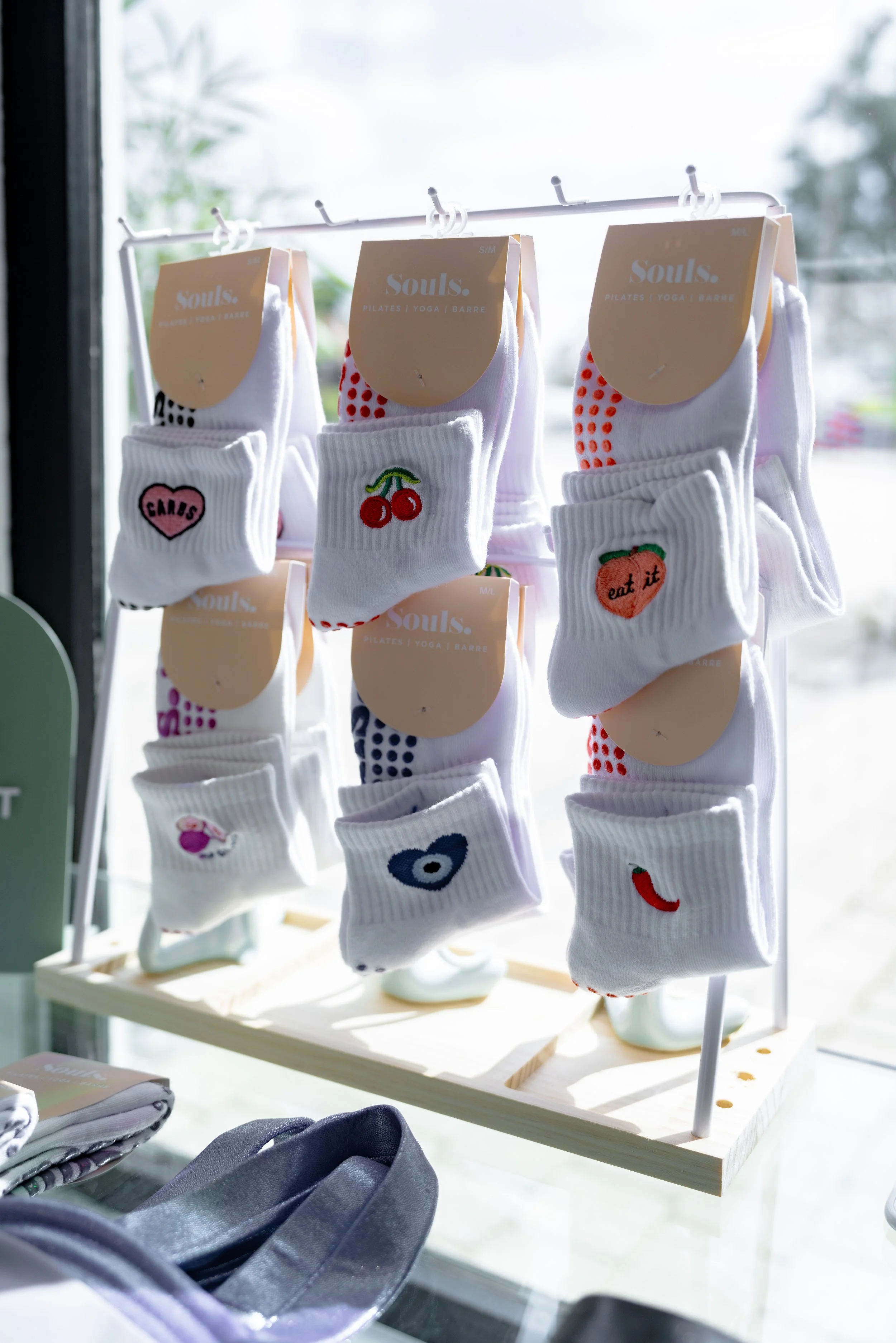 Display of white Souls Grip socks with colorful embroidered patches, including cherries, a heart with 'GAMS,' a peach with 'eat it,' a fish, a chili pepper, and other designs, hanging on a rack in a bright store window.