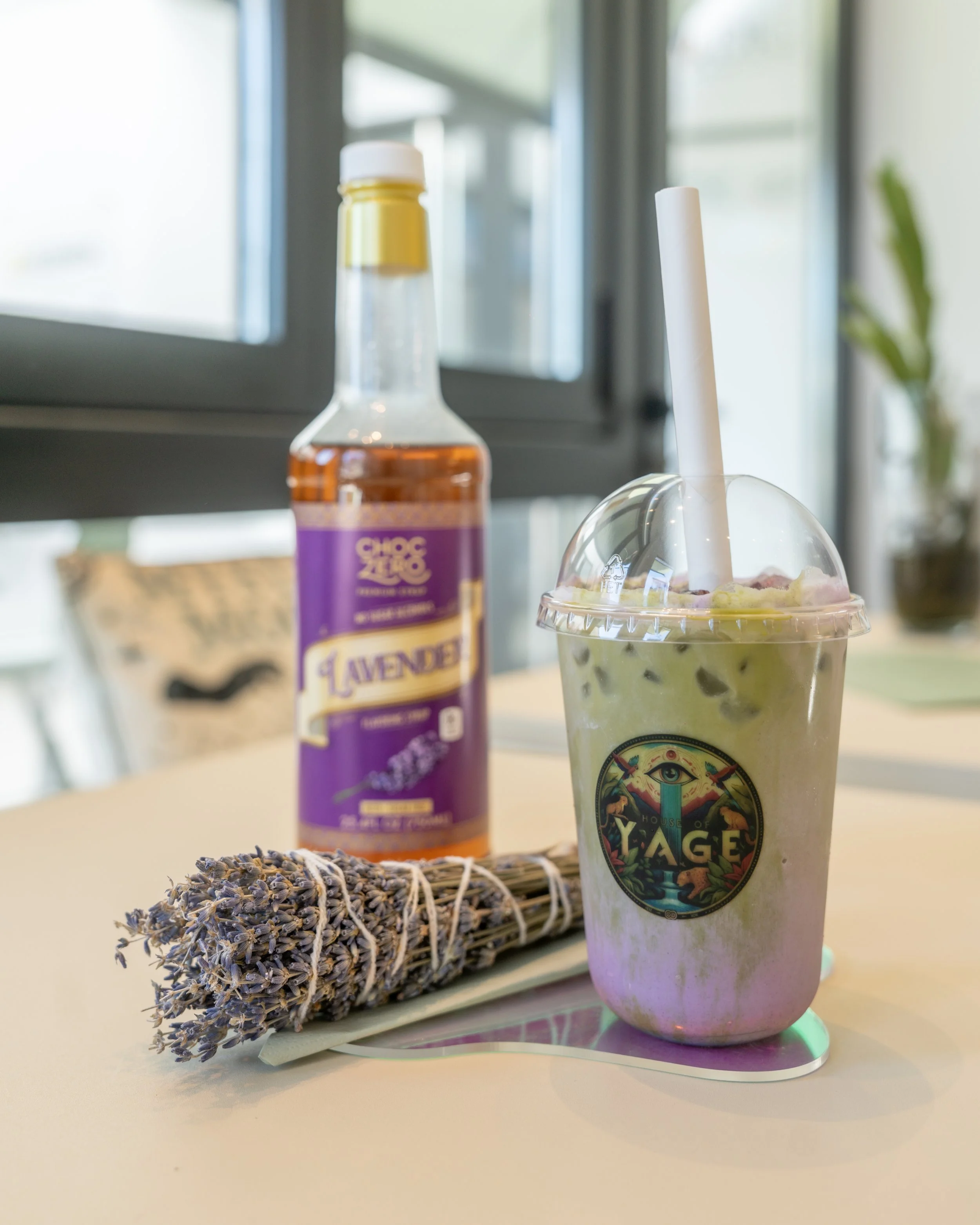 A lavender-infused beverage in a clear plastic cup with a lid and straw, a bottle of lavender syrup, and a bundle of dried lavender on a white table in a bright room with large windows.