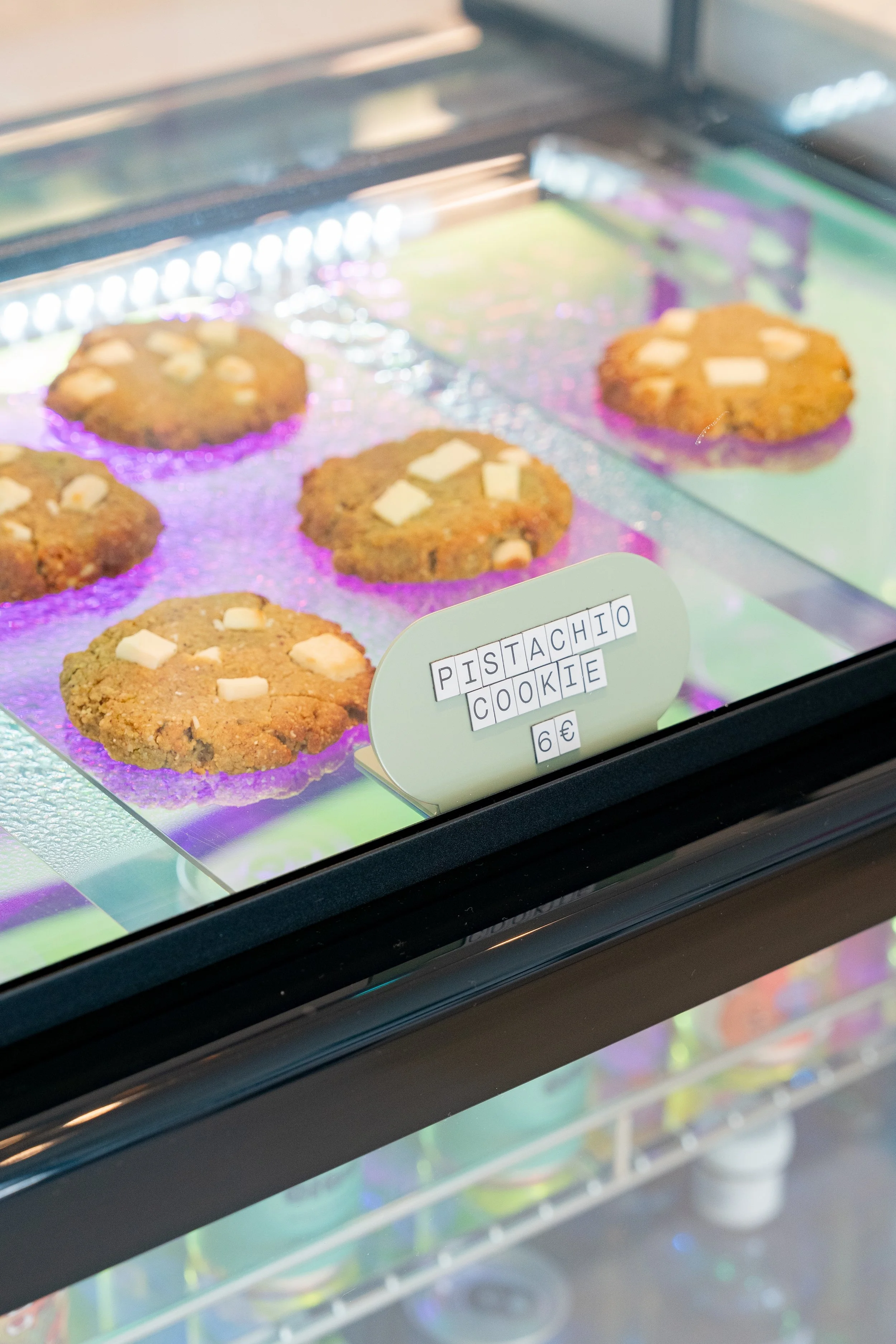 Display case with five pistachio cookies topped with white chocolate chunks, labeled as 'Pistachio Cookie' priced at 6 euros.