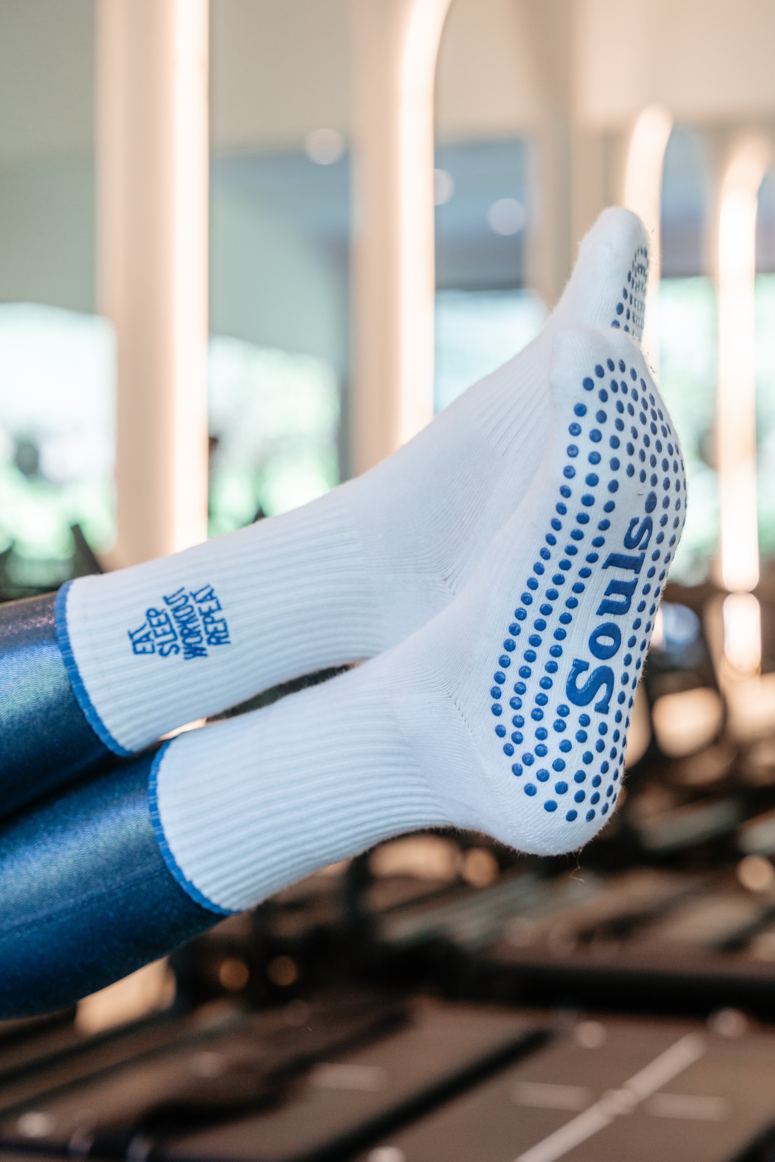 Close-up of a person wearing white Souls Grip socks with blue dots and lettering, resting their feet on gym equipment in a Lagree fitness studio.