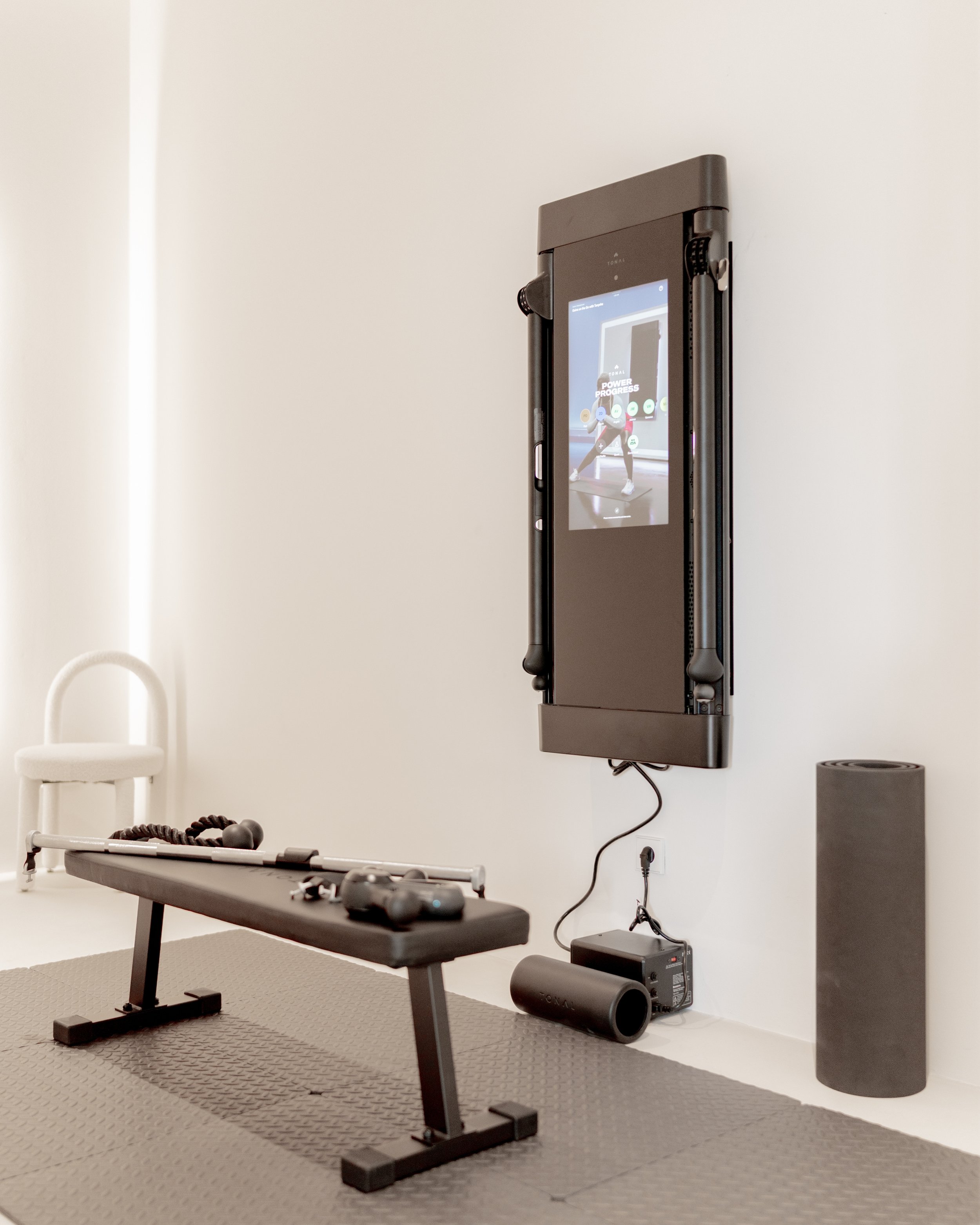 Home fitness room with a wall-mounted screen displaying a fitness class, a bench with workout equipment, and a foam roller on the floor.