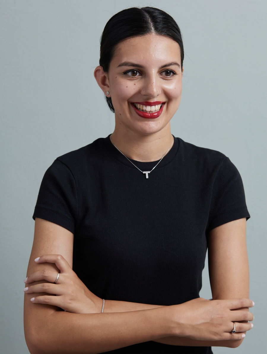 A smiling woman with dark hair pulled back, wearing a black t-shirt, a silver necklace with a T pendant, and small earrings, standing against a plain light-colored background.