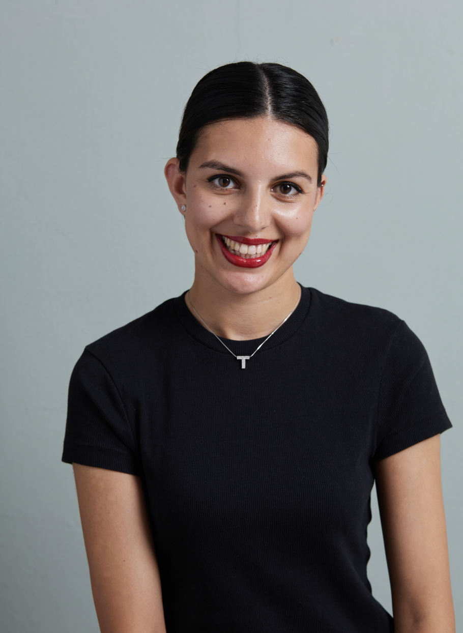A young woman with dark hair pulled back, smiling, wearing a black t-shirt and a silver necklace with a T pendant, standing against a plain light-colored wall.