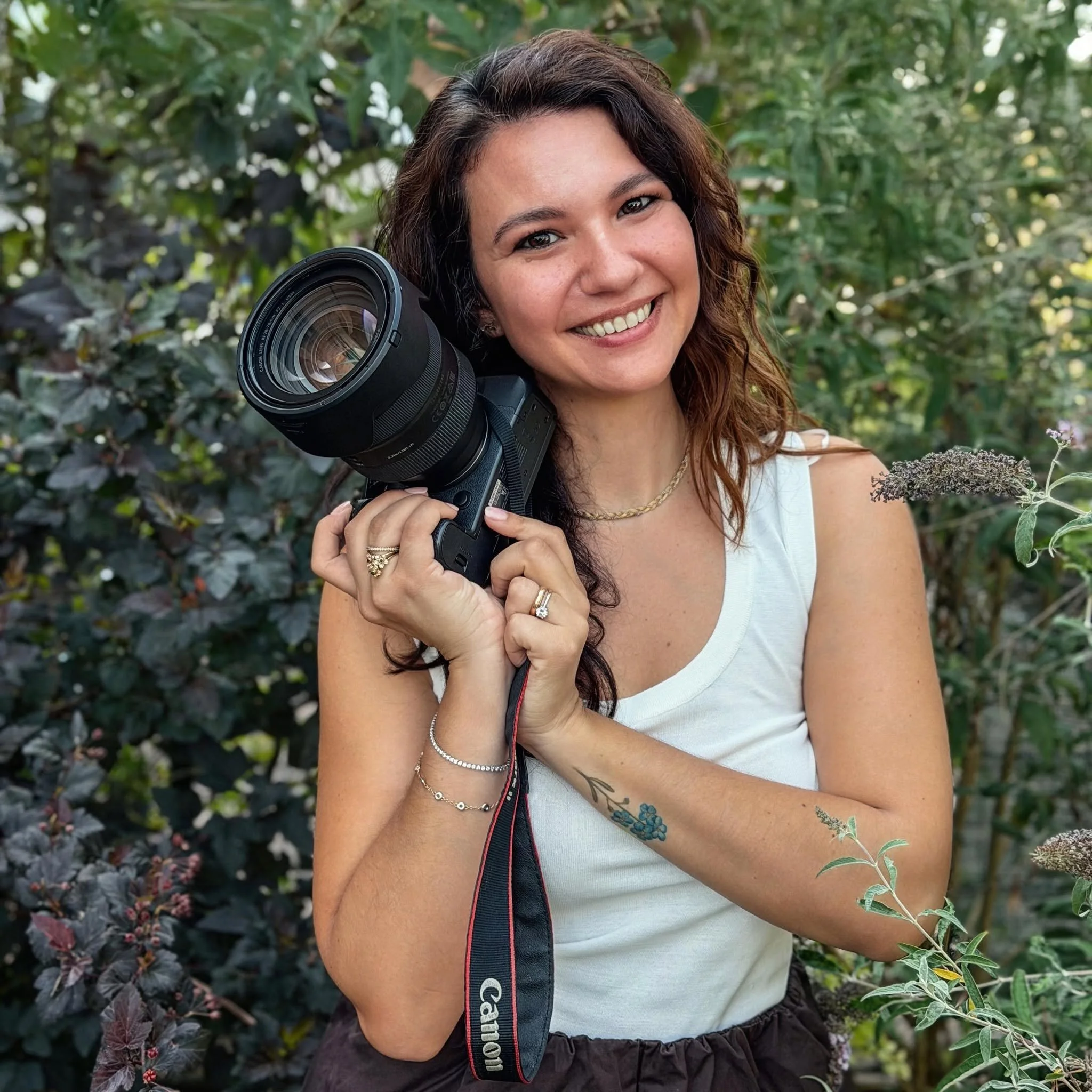 Ragazza sorridente con capelli ricci castani, indossa una maglietta bianca e una collana, tiene una fotocamera Canon con l'obiettivo rivolto verso di sé, si trova in un giardino circondata da piante verdi e fiori.