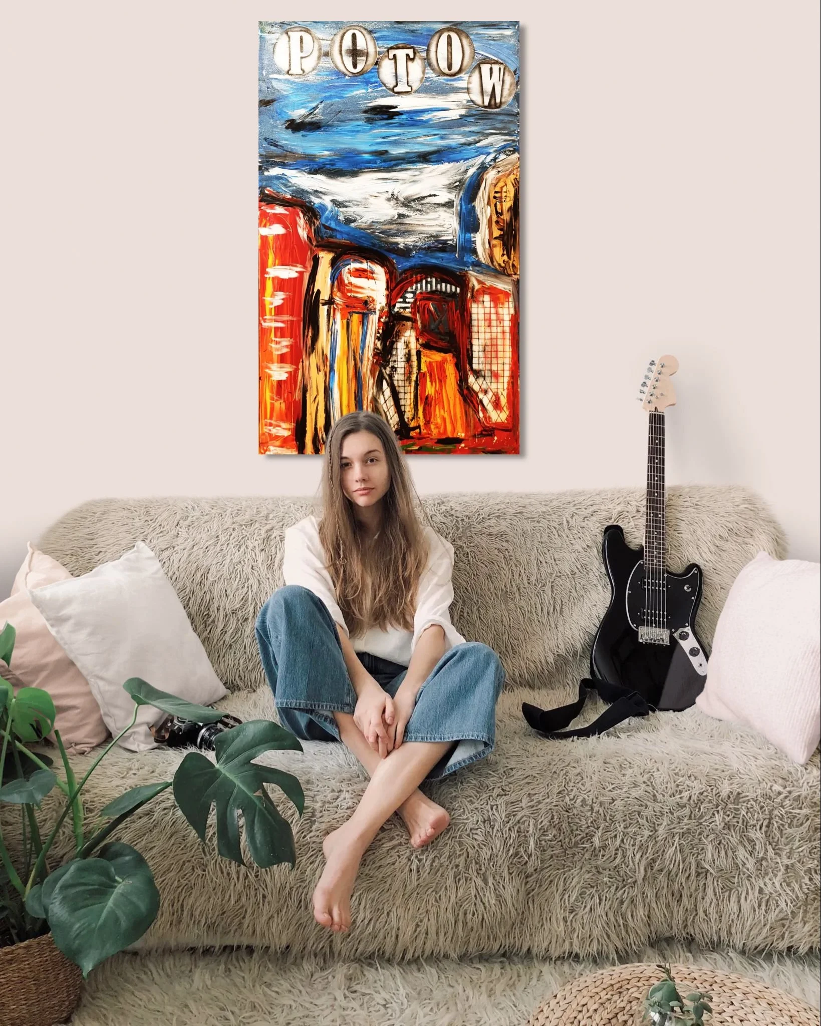 A young woman sitting on a beige furry sofa with pillow cushions, in front of an abstract colorful painting, with an electric guitar placed next to her, in a cozy living room.