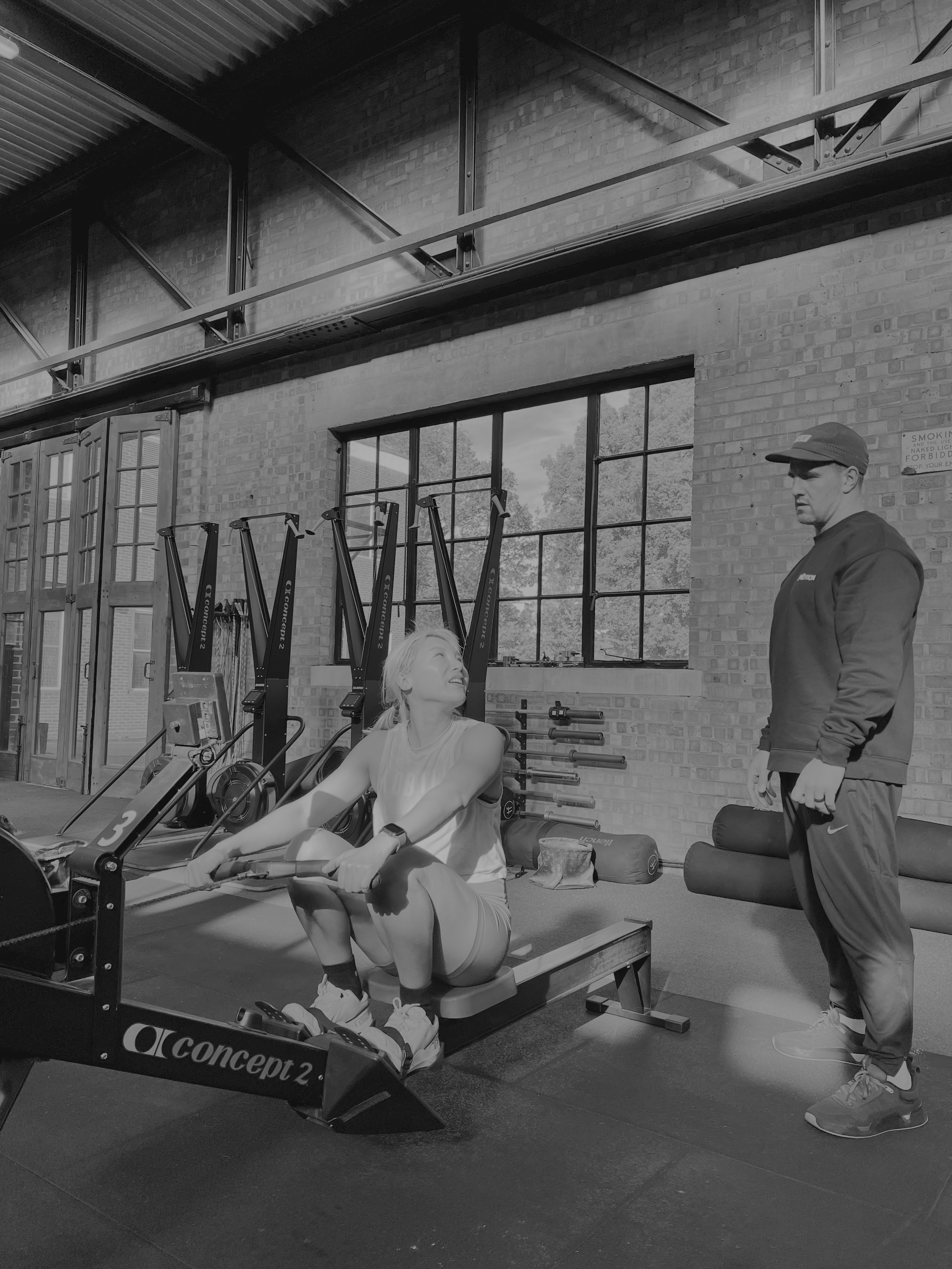 A woman performs rowing exercise on a Concept2 rowing machine in a gym while a trainer stands nearby observing her.