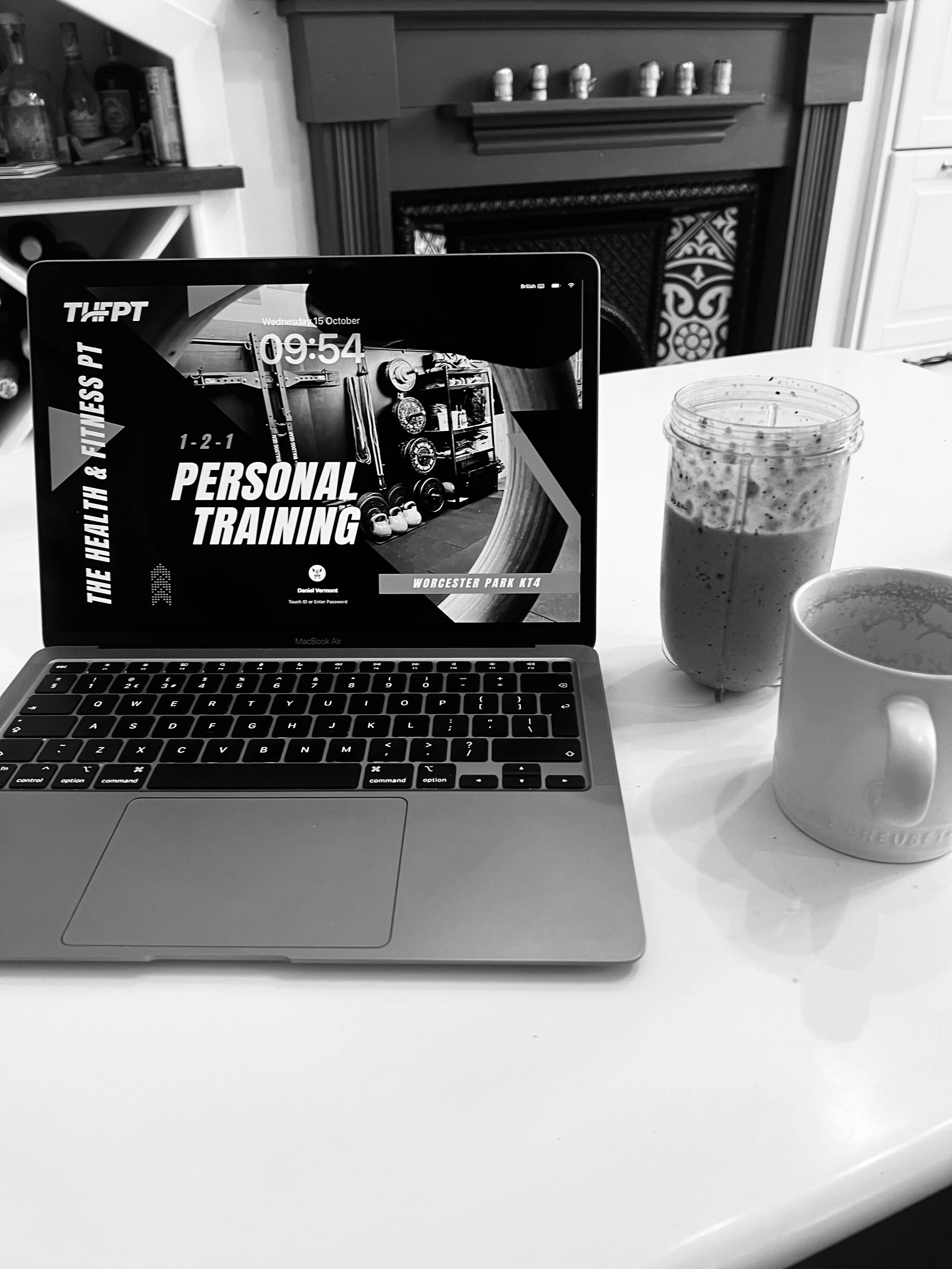 A MacBook Air displaying a fitness class schedule, a protein shake in a tall jar, and a mug on a white table in front of a fireplace.
