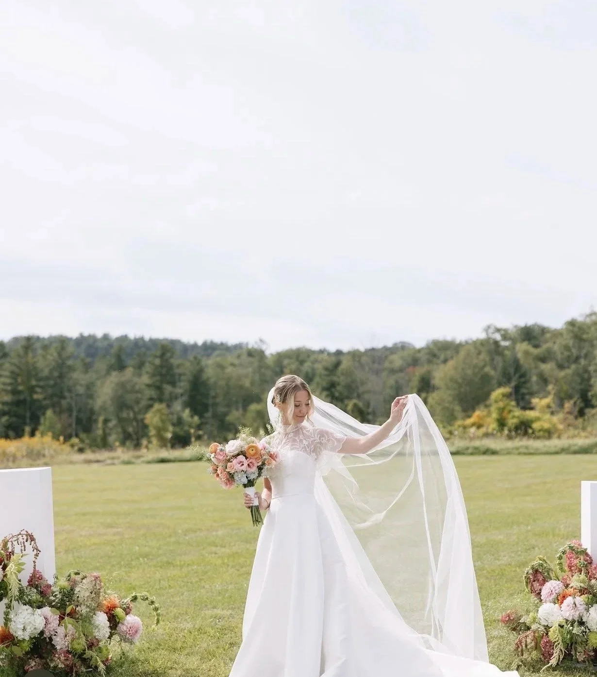 A bride in a white wedding dress standing outdoors in a grassy field, holding a bouquet of pink and white flowers, lifting her veil with one hand, with floral arrangements on either side.