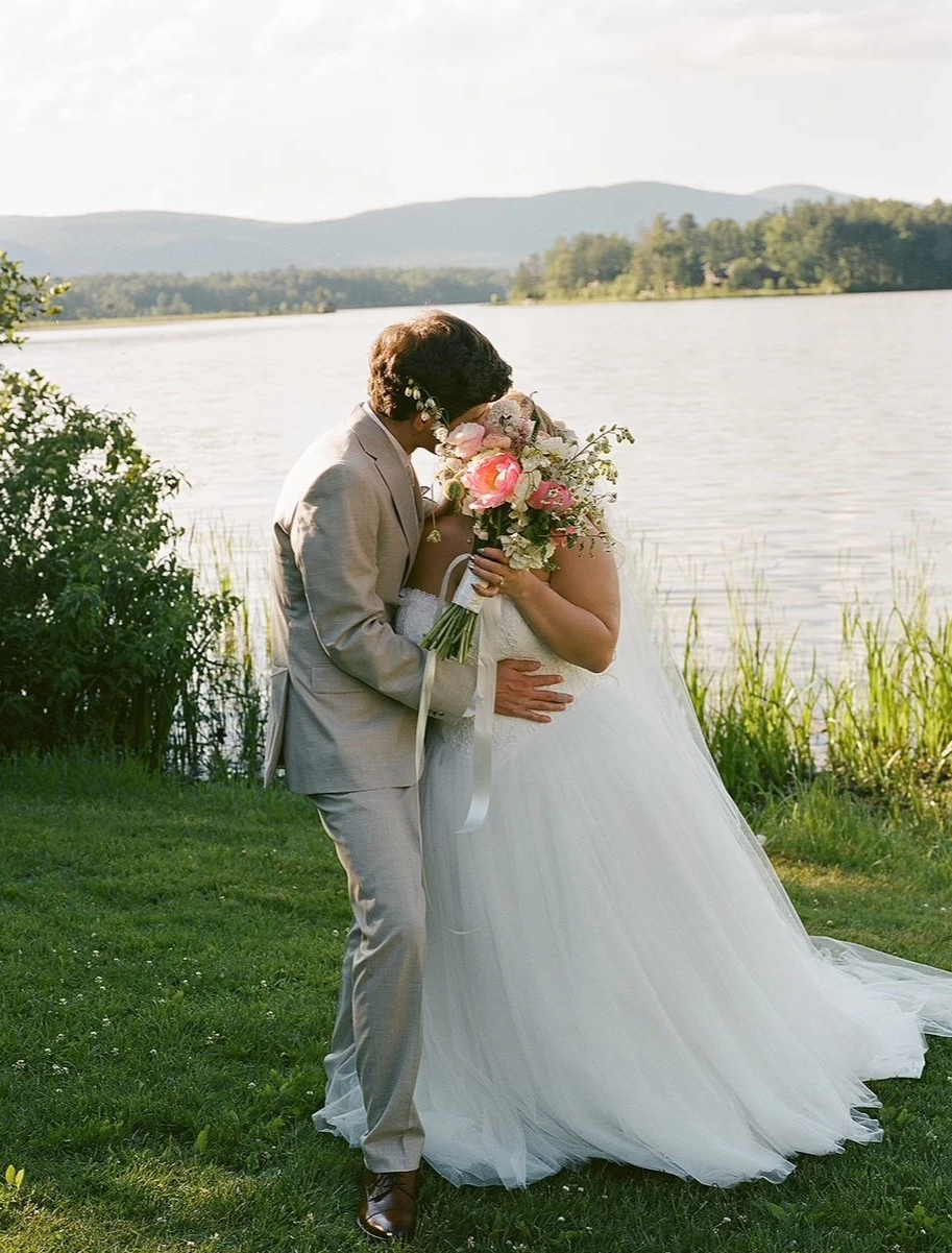A bride and groom standing close near a lake, embracing, with the groom holding a bouquet of pink and white flowers, in a scenic outdoor setting.