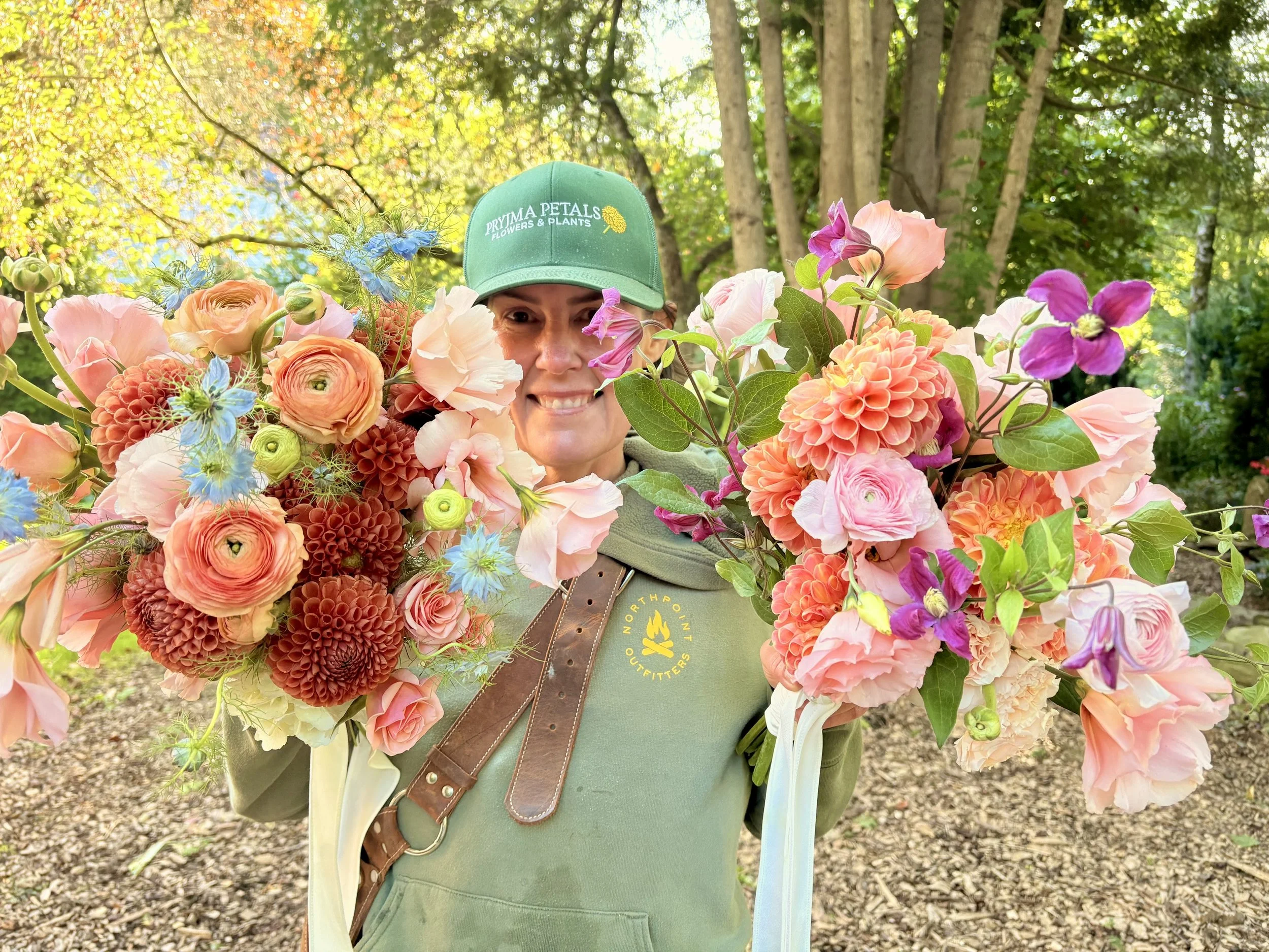 Close-up of a person smiling and holding a large bouquet of colorful flowers, wearing a green cap with green trees and sunlight in the background.