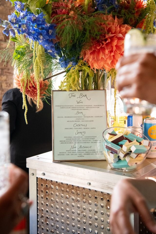 A bouquet of colorful flowers including blue, red, and orange blooms in a tall glass vase on a bar counter, with a drinks menu in a frame and candies in a bowl nearby.