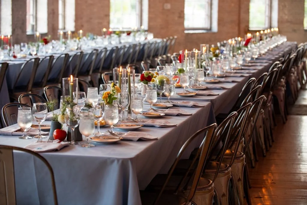 Long banquet table set with white tablecloth, glasses, plates, napkins, and floral centerpieces with candles, in a brick-walled room with windows.