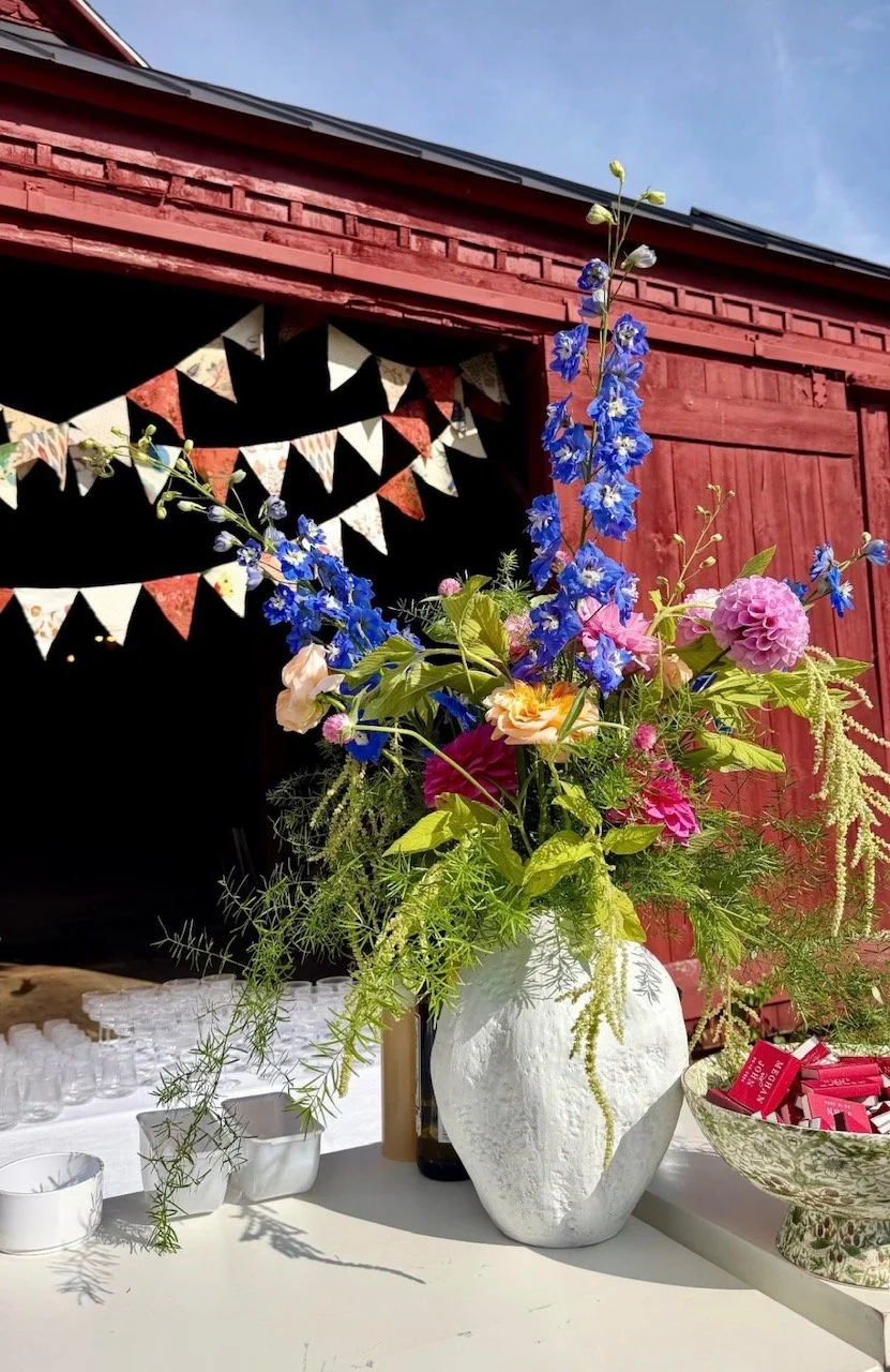 A vibrant bouquet of pink, purple, and blue flowers in a white textured ceramic vase, set against a red wooden building with bunting decorations on the black interior background.
