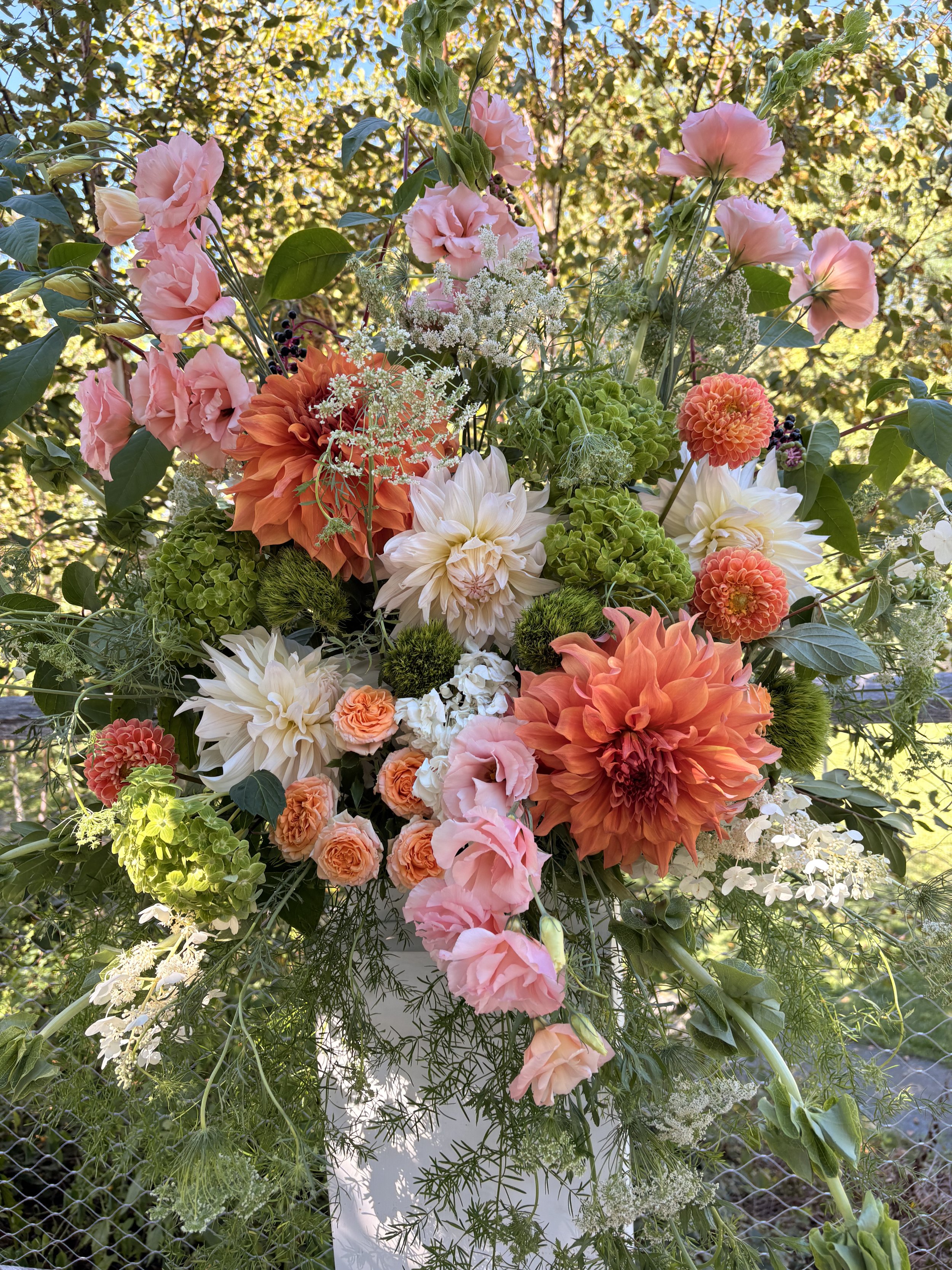A colorful flower arrangement with pink, orange, white, and green flowers and foliage, arranged outdoors against a background of trees and blue sky.