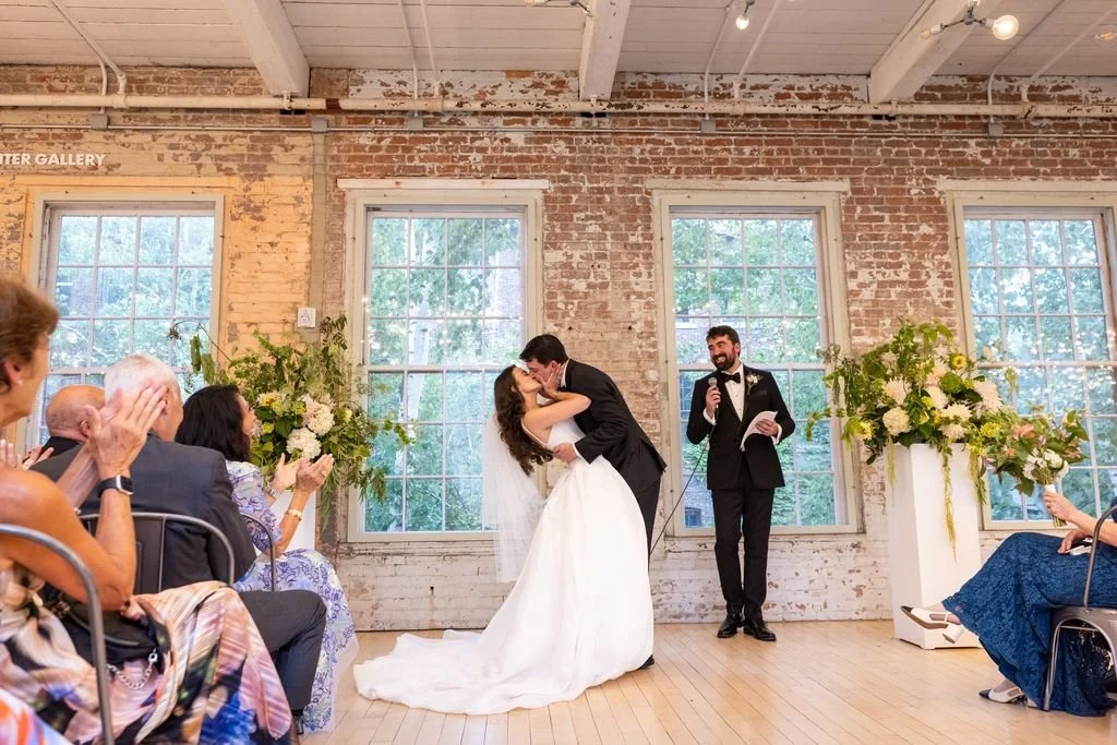 A bride and groom kiss during their wedding ceremony in a rustic room with exposed brick walls and large windows. Guests are clapping and smiling while the officiant reads.
