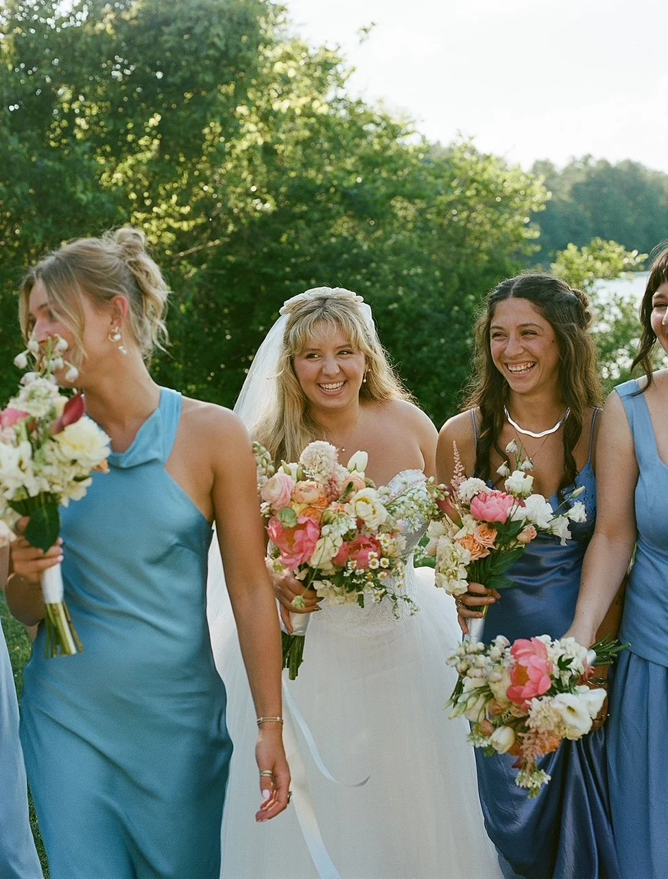 Bride and bridesmaids holding bouquets outdoors, with green trees and sky in the background, during a wedding celebration.