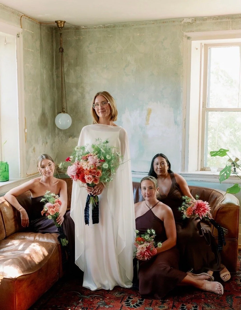 Four women, dressed in bridesmaid dresses and a bride in a white gown, pose with bouquets in a vintage living room with weathered walls, large window, and brown leather sofa.