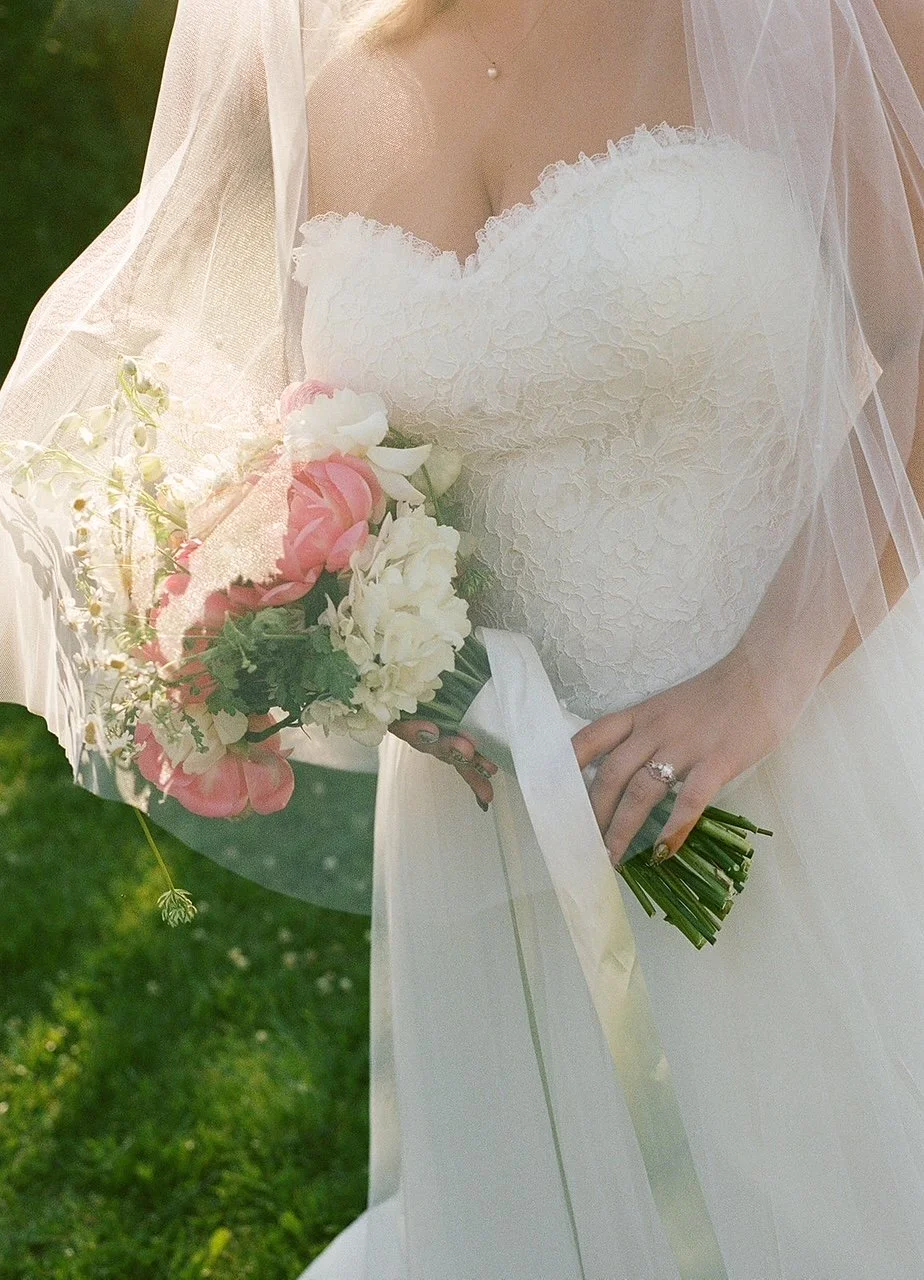 Close-up of a bride holding a bouquet of pink and white flowers, wearing a wedding dress and veil, outdoors on grass.