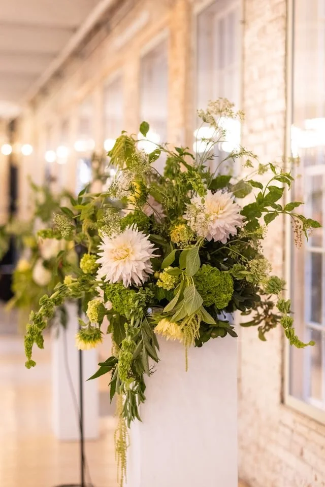 Fresh flower arrangement with white dahlias, green hydrangeas, and assorted greenery in a white vase, set against a rustic brick wall in a bright indoor space.