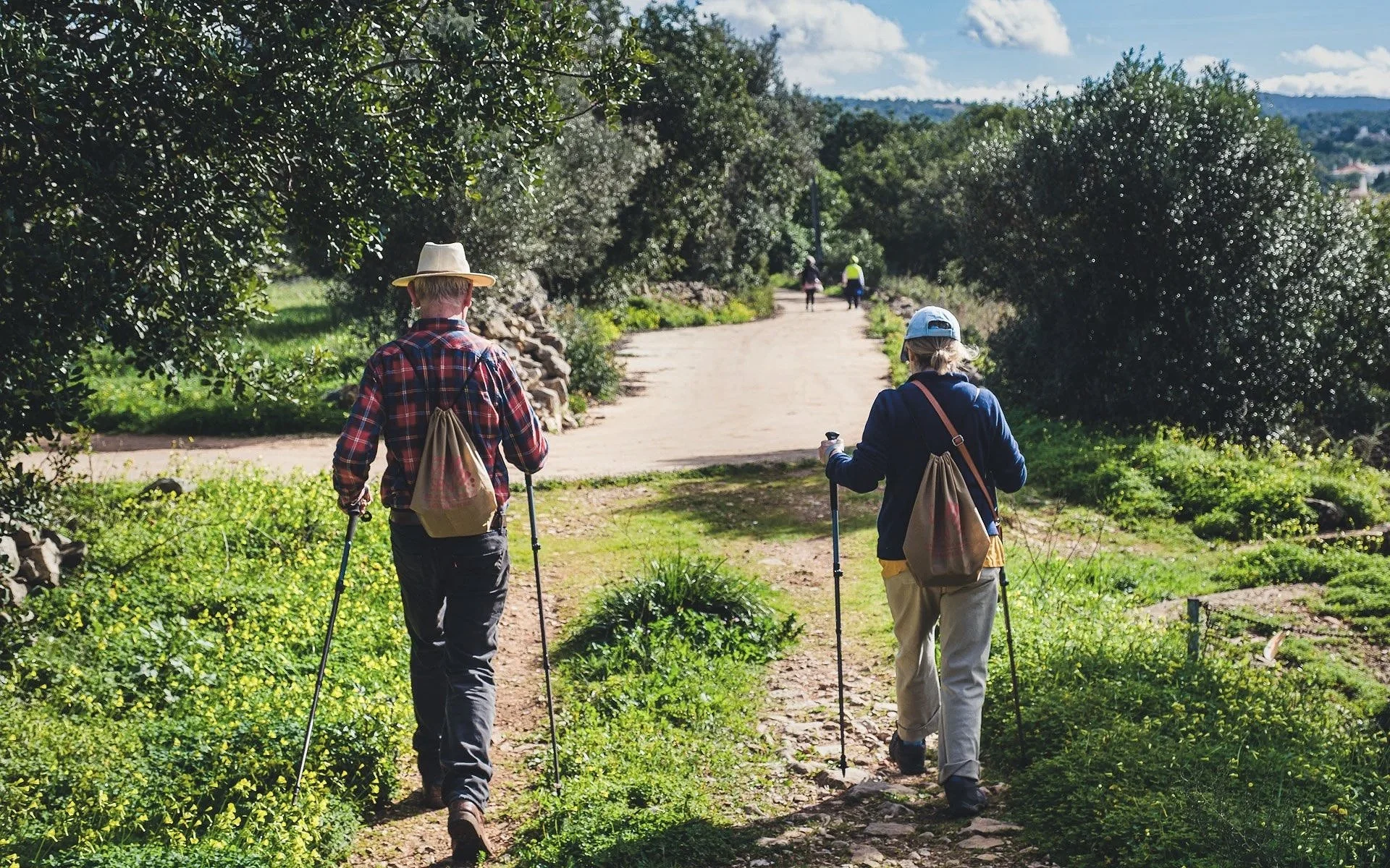 Homem a caminhar à saída de um trilho, com dois bastões, chapéu e mochila