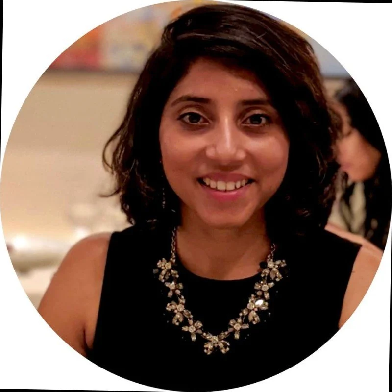 Smiling woman with short dark hair wearing a black top and a decorative necklace in a softly focused indoor setting.