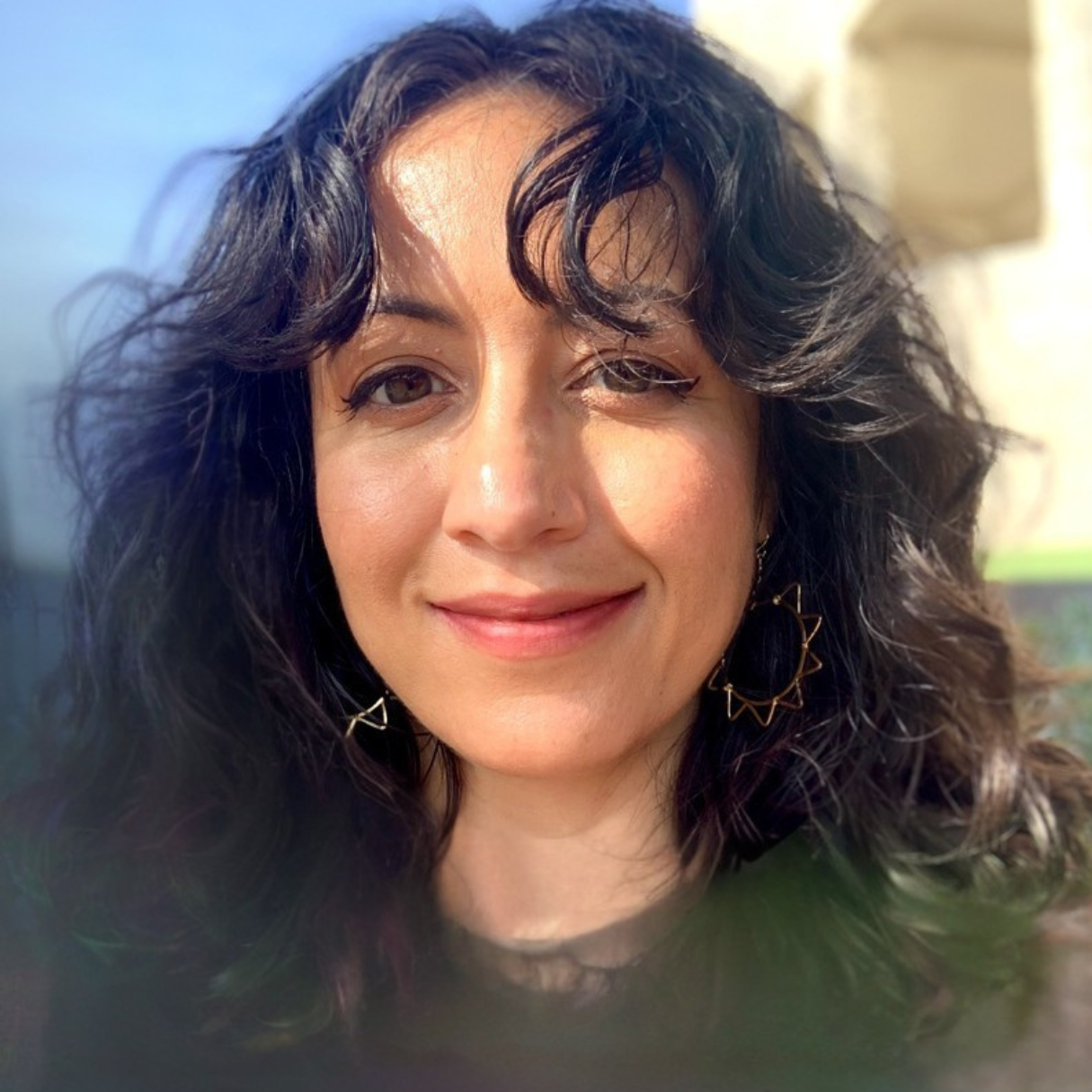 A woman with long, wavy dark hair is smiling outdoors, wearing star-shaped earrings, with sunlight illuminating her face. The background is slightly blurred.