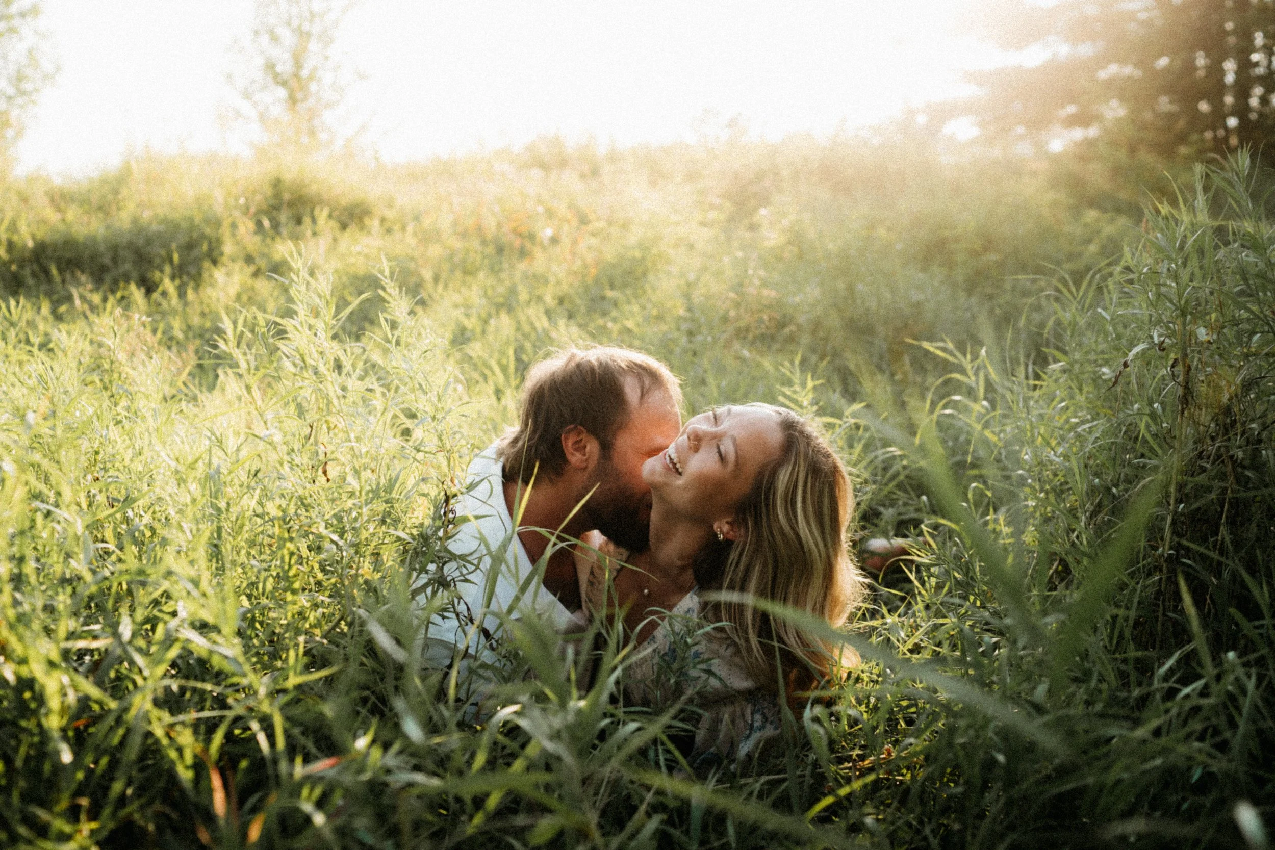 A couple lying in a grassy field, touching faces and smiling at each other, with sunlight in the background.