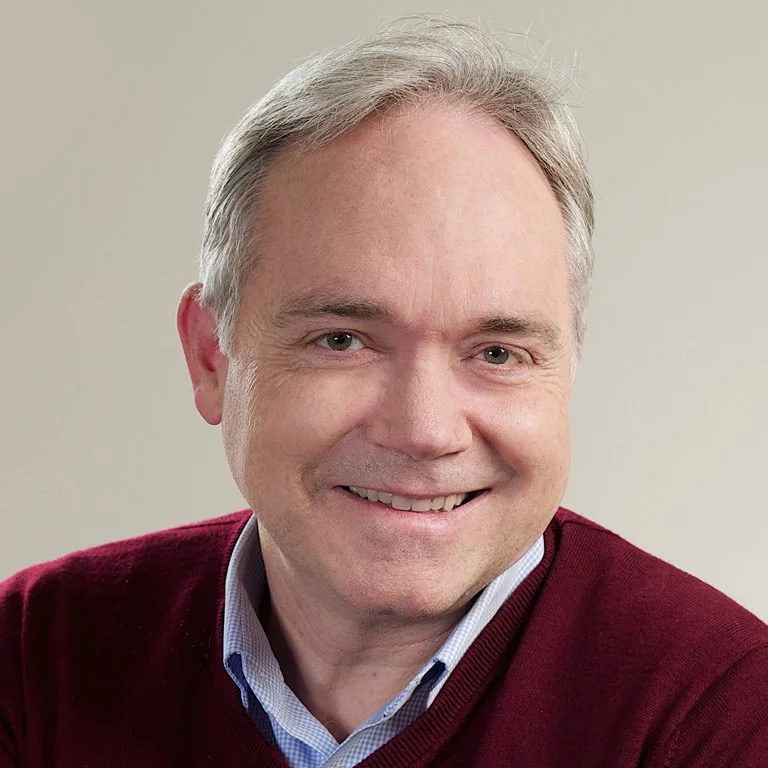 Portrait of a smiling middle-aged man with gray hair, wearing a burgundy sweater over a collared shirt.