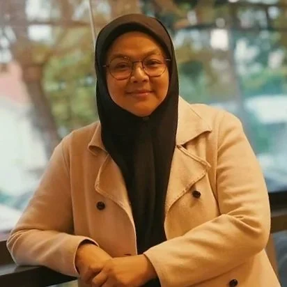A woman wearing glasses and a black hijab, smiling and sitting in an indoor setting with a window in the background.