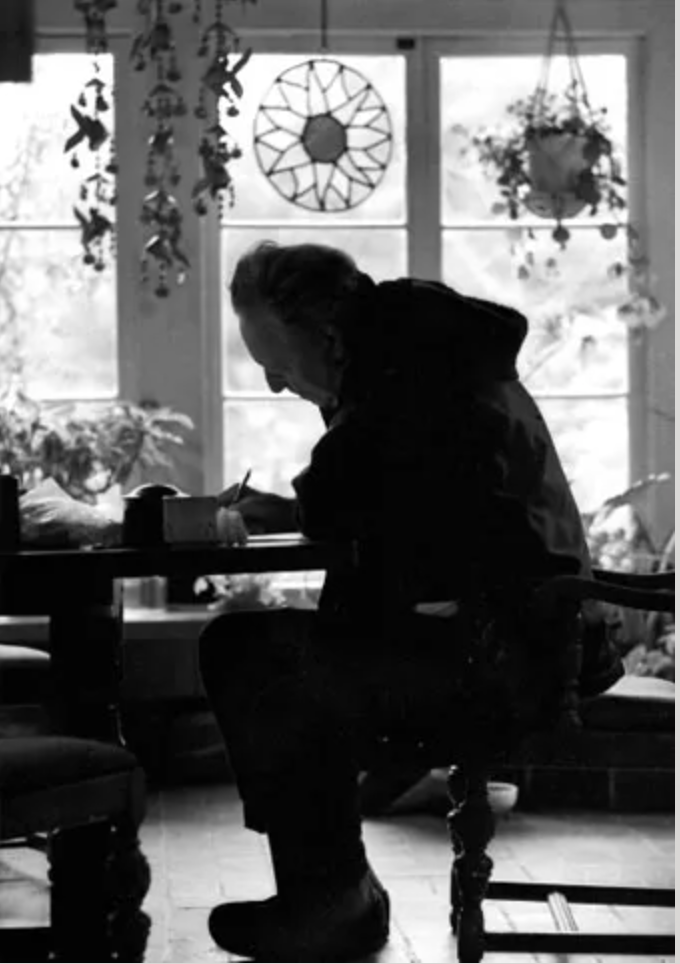 Gregory Bateson sitting at a table in a room with large windows, writing or drawing at the table, with plants hanging and on the windowsill in the background.