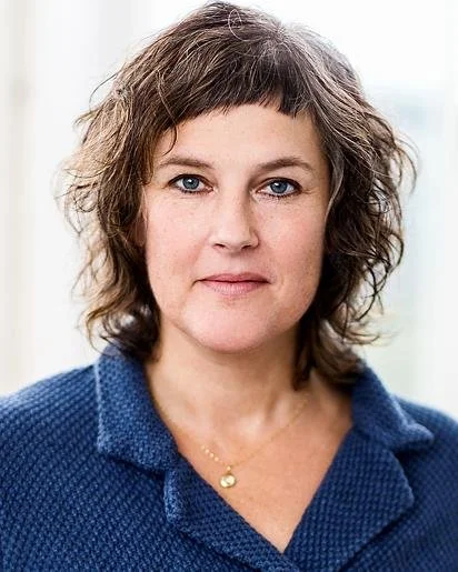 A woman with short, curly brown hair and blue eyes, wearing a blue textured shirt and a gold necklace, standing indoors with a soft background.