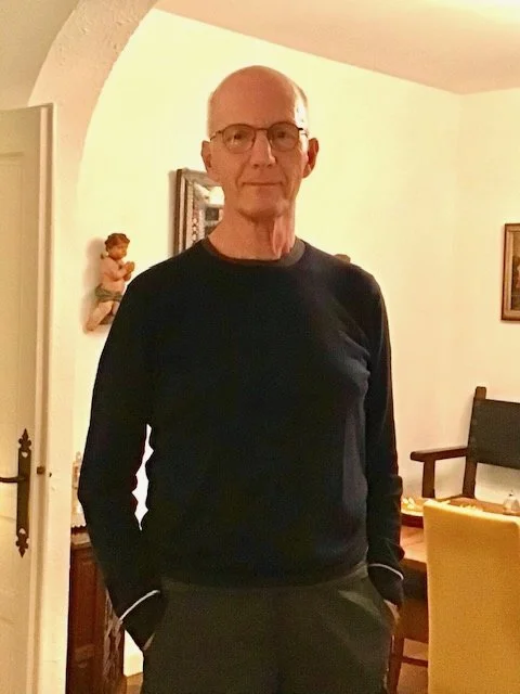 An older man with glasses and a black sweater standing in a dining room with his hands in his pockets.
