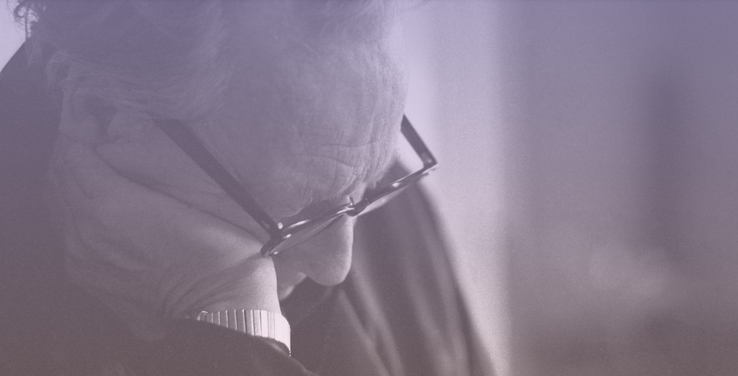 Close-up of Gregory Bateson wearing glasses, looking downward, with a soft light illuminating their face, and a blurred background.