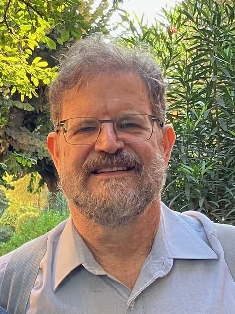 A smiling middle-aged man with glasses, a beard, and gray hair, outdoors among green trees and plants.