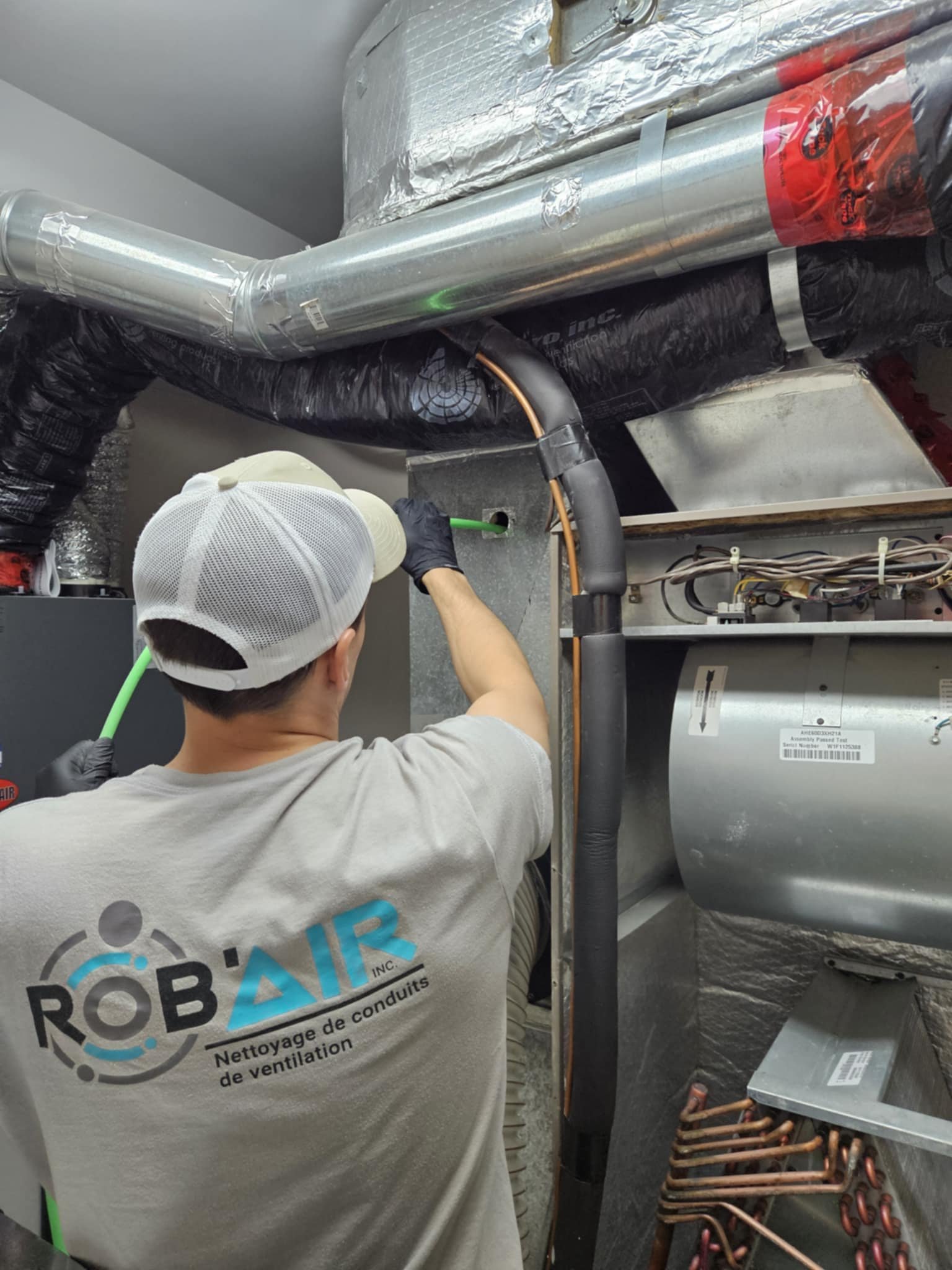 Technician working on HVAC or ventilation system, wearing a gray t-shirt with 'ROB'AIR' logo and text in French, holding a green tube.