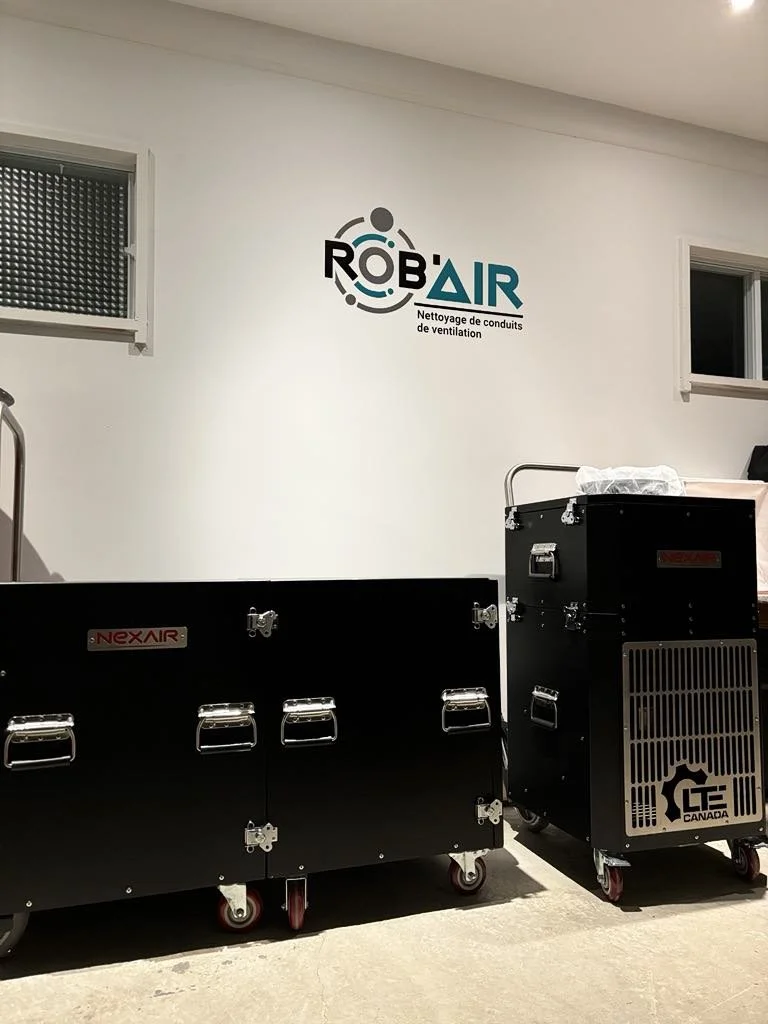 Two large black industrial ventilation cleaning machines with caster wheels positioned on a concrete floor in front of a white wall with a logo that reads "ROBAIR Nettoyage de conduits de ventilation". One machine has a metal vent on the front labeled "GT Canada".