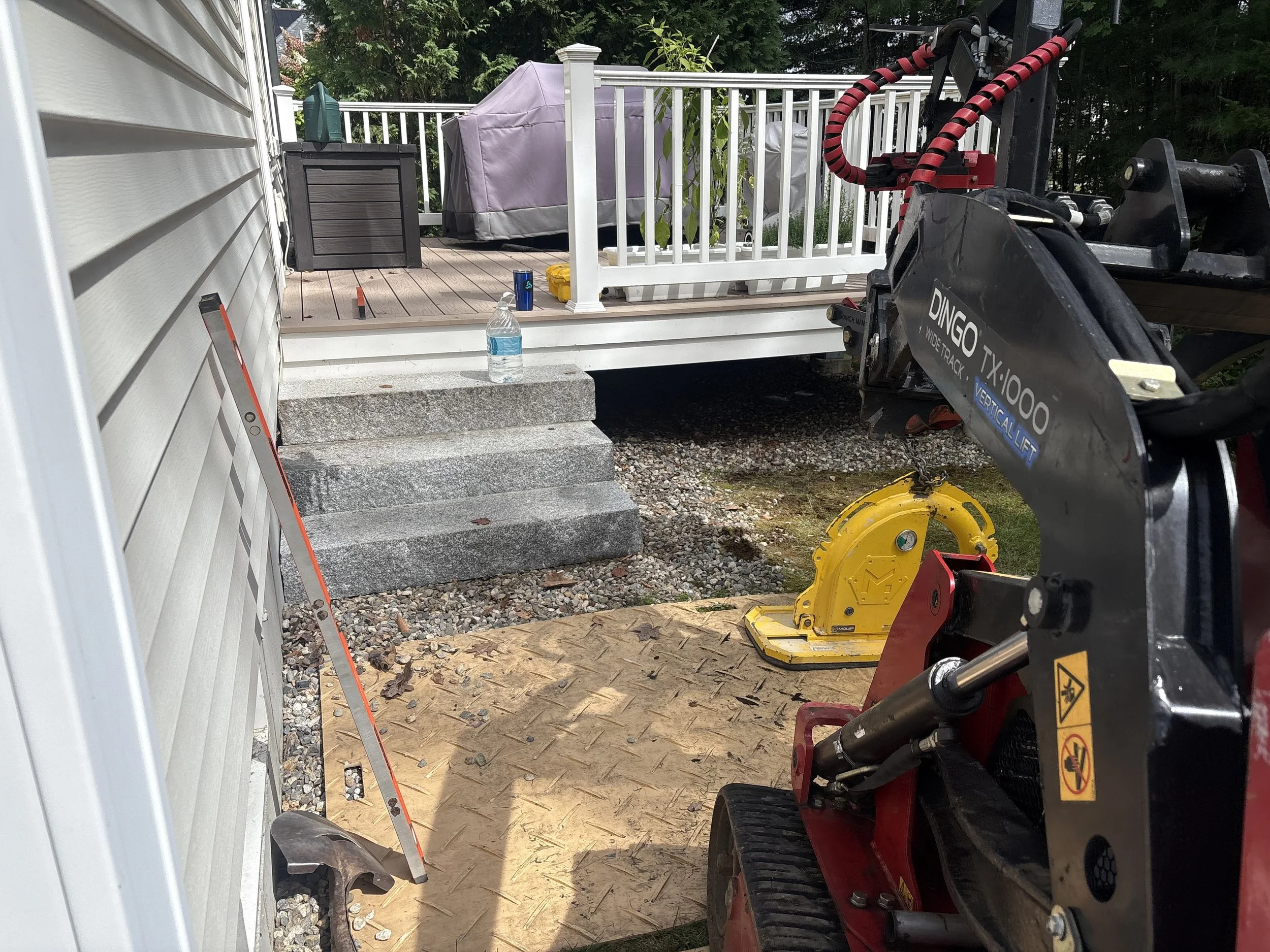 View from inside a construction site showing steps leading to a porch, with various tools and materials including a water bottle, a level, and a small excavator, during daytime.