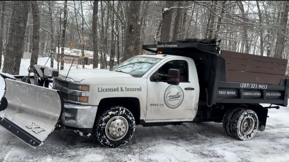 A white pickup truck with a snow plow attached to the front, parked on a snow-covered road in a wooded area. The truck has signage for Cassella Enterprises, offering tree work, landscaping, and excavation services.