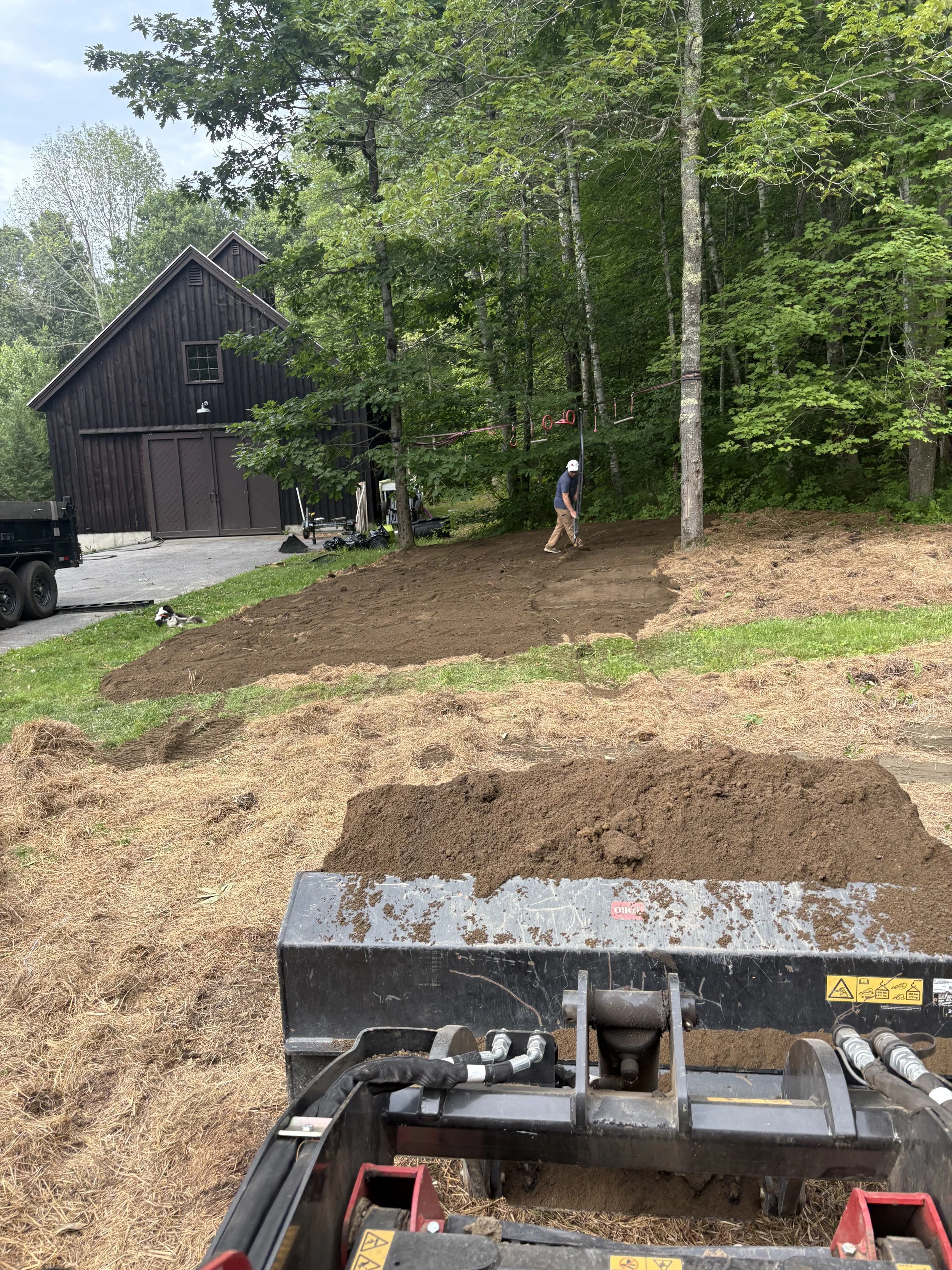 View from the front of a tractor or bulldozer with a metal blade, pushing dirt in a landscape with trees and a wooden barn. A person is in the background near trees, and the tractor's hydraulic parts are visible in the foreground.